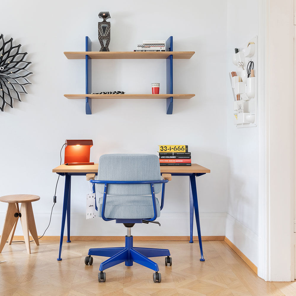 Modern eingerichtetes Arbeitszimmer mit hellblauem Schreibtischstuhl und Schreibtisch mit blauen Beinen. Auf dem Tisch steht die rote Tischlampe »Lampe de Bureau« von VITRA, daneben Bücher und ein kleiner Holzhocker. Wandregale, Wanddeko und Organizer ergänzen die Szenerie.