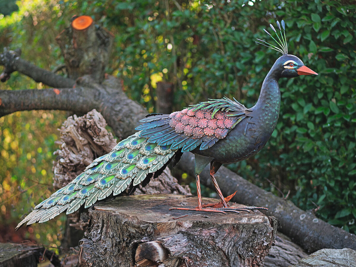 Metall-Gartenfigur eines kunstvoll gestalteten Pfaus, sitzend auf einem Baumstamm in einem üppigen Garten