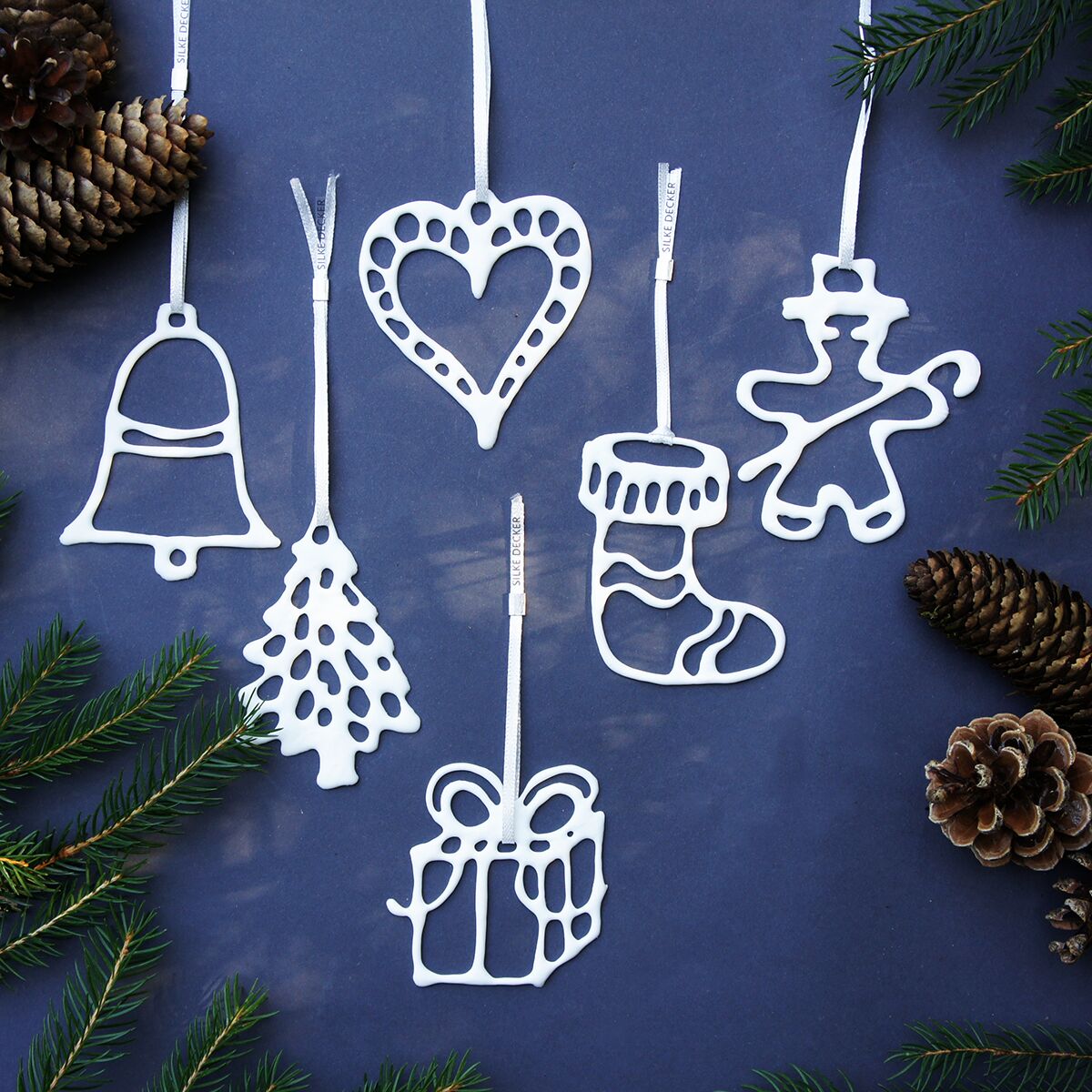 Sechs filigrane, handgefertigte Weihnachtsanhänger aus weißem Porzellan liegen auf dunkelblauem Hintergrund. Dargestellt sind Motive wie Glocke, Herz, Tannenbaum, Geschenk, Stiefel und Lebkuchenfigur. Kiefernzweige und Tannenzapfen schmücken die Szenerie winterlich.