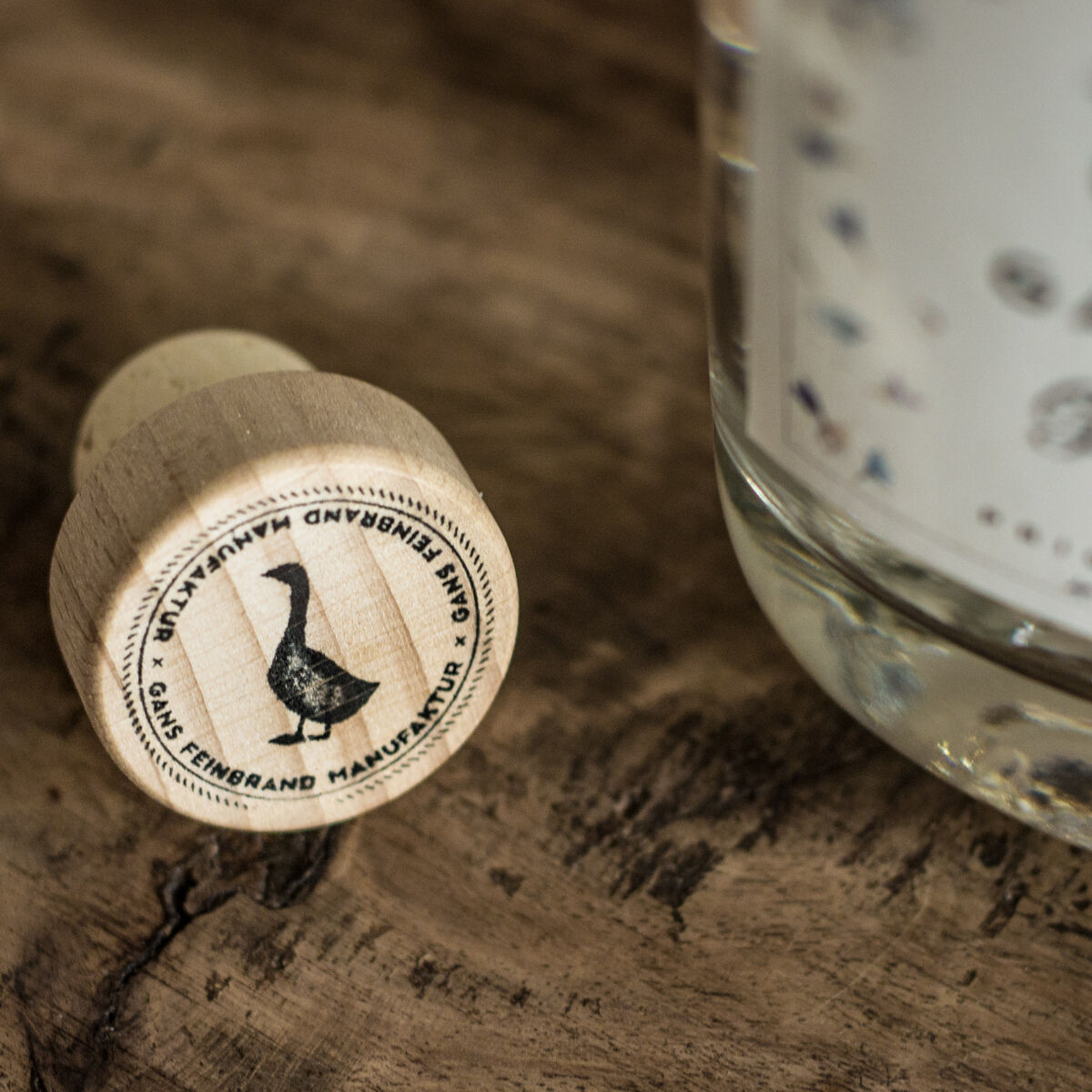 Eine Nahaufnahme eines Holzkorkens mit Gans-Emblem und einer Gin-Flasche der Marke »Gin« von GANS auf einer hölzernen Oberfläche.
