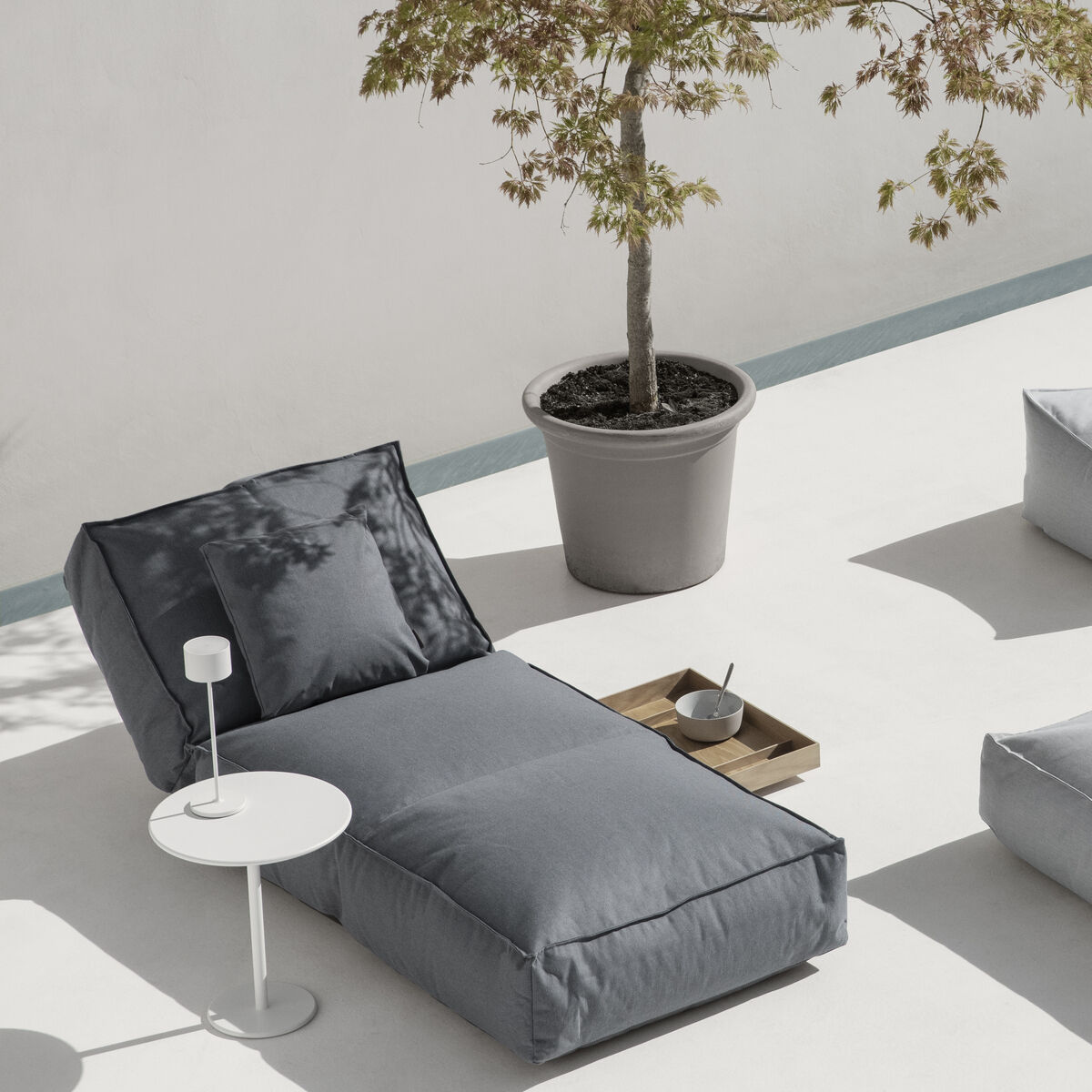 Ein moderner Wohnbereich im Freien mit minimalistischer Einrichtung und neutralen Farben. Weiße und graue Möbelstücke sind symmetrisch arrangiert, und ein Baum in einem grauen Topf steht zur Dekoration. Eine Mobile LED-Tischleuchte »FAROL« ist auf einem Beistelltisch neben einer Sonnenliege platziert.