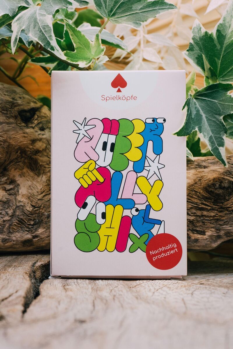 Die Vorderseite einer Verpackung des Kartenspiels 'Queer Allyship' von Spielköpfe ist zu sehen. Der Hintergrund ist aus hellem, naturbelassenem Holz mit grünen Blättern am Rand. Das Design der Verpackung ist bunt mit auffälligen, handgezeichneten Buchstaben in Weiß, Grün, Blau, Gelb, Rot und Violett. Ein roter Aufkleber unten rechts zeigt 'Nachhaltig produziert'.