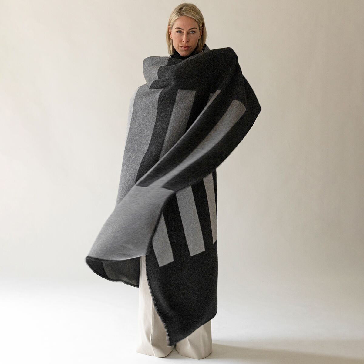 Eine Frau steht vor hellem, neutralem Hintergrund und ist in eine großformatige, anthrazitfarbene Kaschmirdecke mit markanten, hellgrauen Streifen gehüllt. Die Decke wirkt weich und hochwertig, ihr edles Design vermittelt Wärme und Komfort.