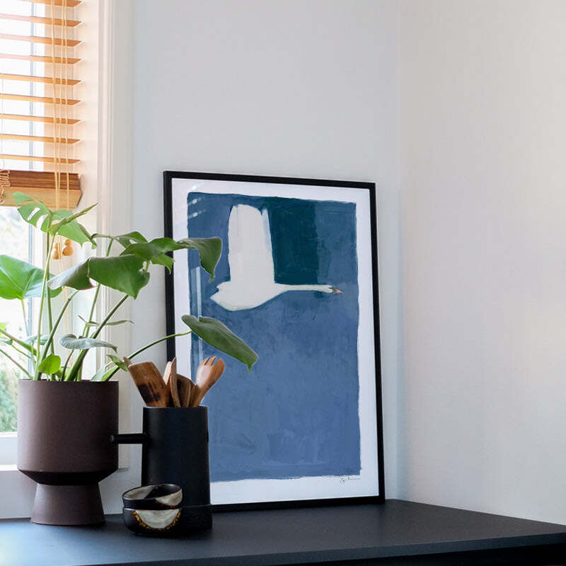 Minimalistisches Poster »The Swan« von Jörgen Hansson in einem schwarzen Rahmen, zeigt einen abstrakt gestalteten weißen Schwan auf blauem Hintergrund. Das Bild steht auf einem modernen schwarzen Sideboard neben einer grünen Zimmerpflanze und dekorativen Accessoires in einem hellen Raum.