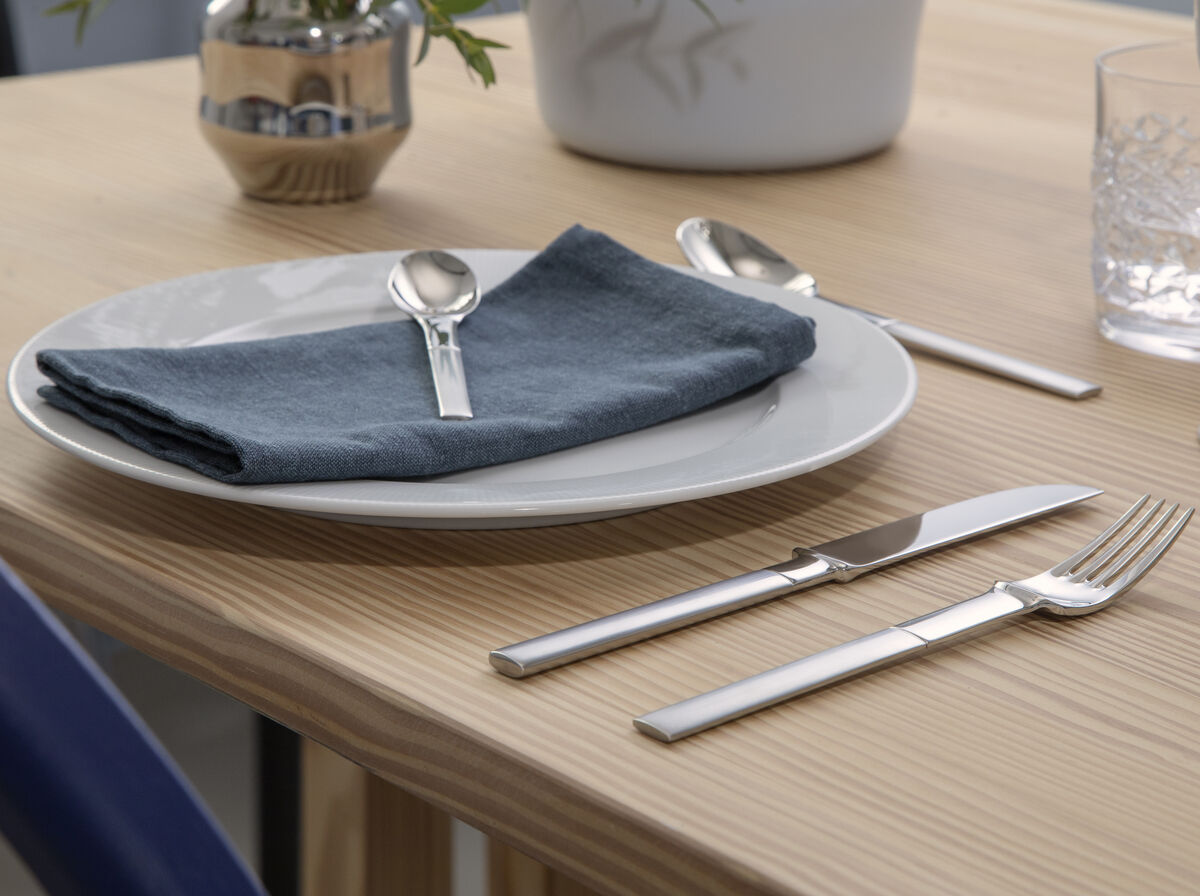 Ein moderner gedeckter Tisch mit einem Besteckset aus Edelstahl, das neben einem weißen Teller mit einer blauen Serviette arrangiert ist. Im Hintergrund sind dekorative Elemente wie Vasen und ein Glas zu sehen.