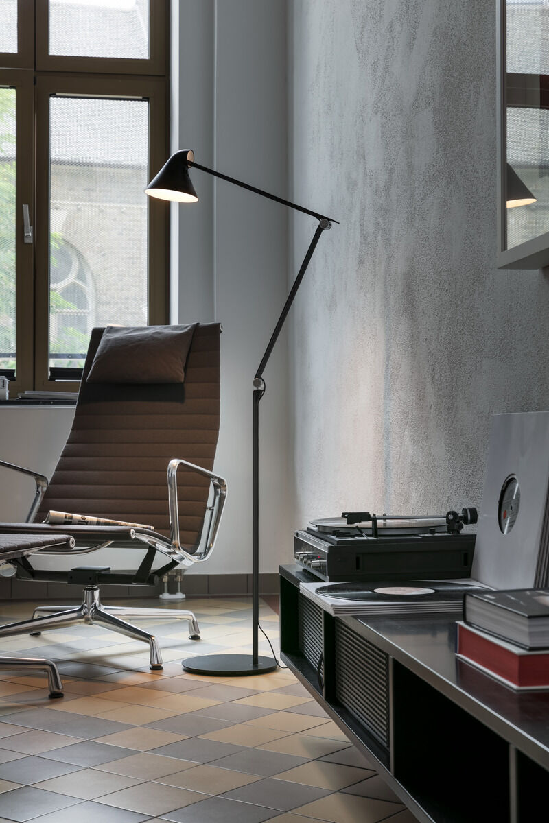 Eine schwarze Stehleuchte der Marke »NJP« steht neben einem braunen Bürostuhl vor einem großen Fenster. Die Lampe hat ein minimalistisches, modernes Design und beleuchtet einen gehobenen Raum mit Schallplattenspieler und dekorativen Accessoires.