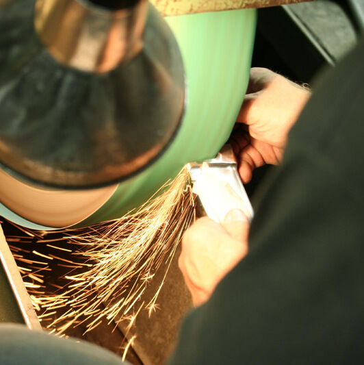 Eine Person bearbeitet ein metallisches Werkzeug an einer Schleifmaschine, wobei Funken sprühen. Das Werkzeug scheint ein Messer oder ein Küchenutensil zu sein, möglicherweise ein Käsemesser mit einem Griff aus Olivenholz.