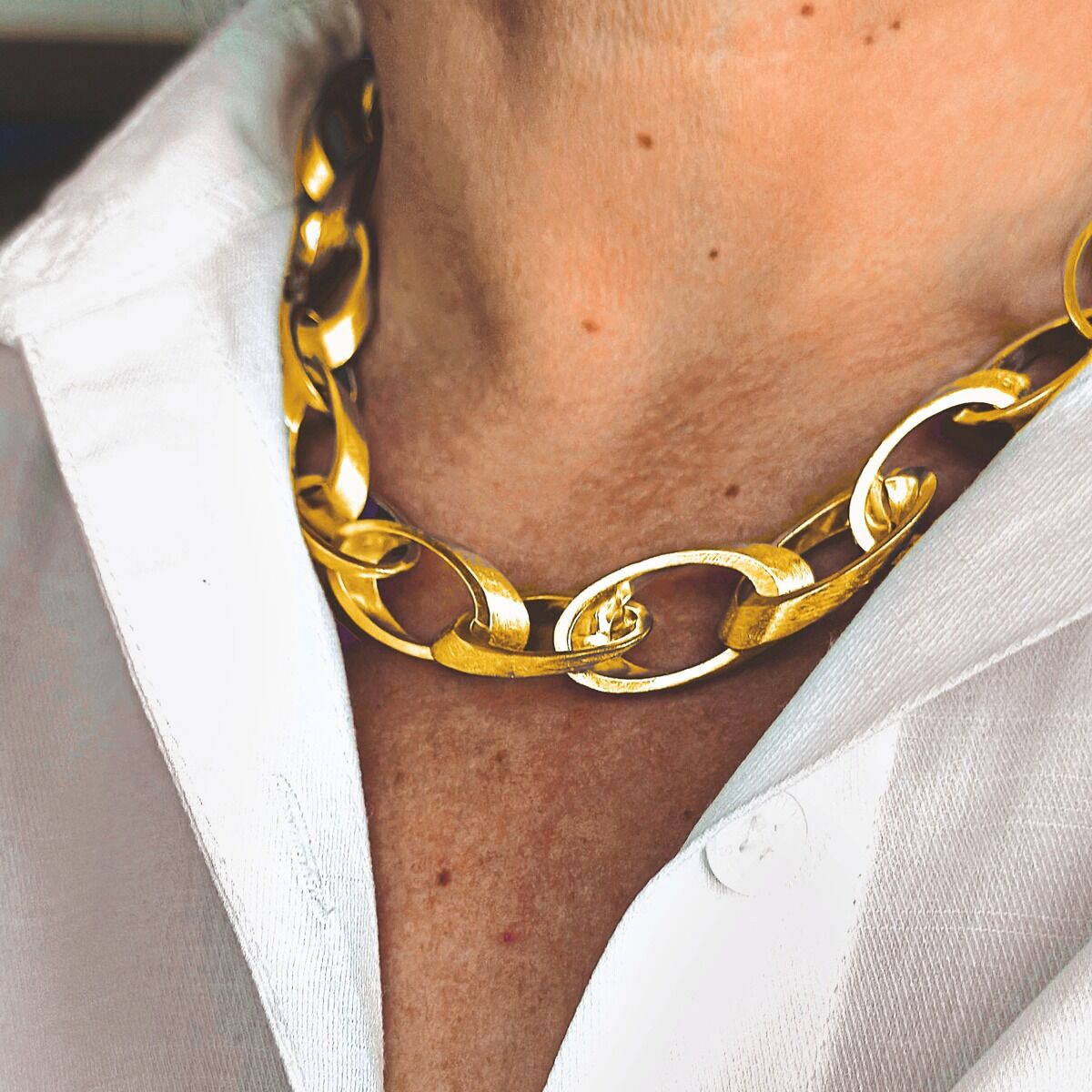 Eine Nahaufnahme zeigt den Hals einer Person mit einem auffälligen, feingoldplattierten Collier aus 925er Silber. Die Kette besteht aus großen, glänzenden Gliedern. Die Person trägt ein weißes Hemd. Das Schmuckstück ist elegant und hochwertig verarbeitet.