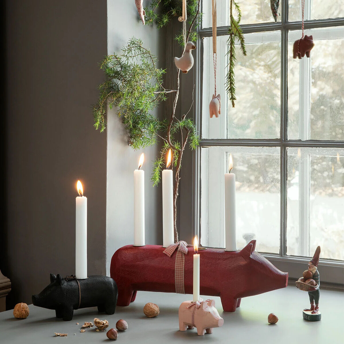 Ein roter Kerzenhalter in Form eines Holz-Schweins von Maileg steht im Mittelpunkt auf einer Fensterbank, dekoriert mit brennenden weißen Kerzen. Daneben stehen kleinere Schweinefiguren, Nüsse und Tannenzweige schmücken das Fenster. Winterliche, gemütliche Atmosphäre.