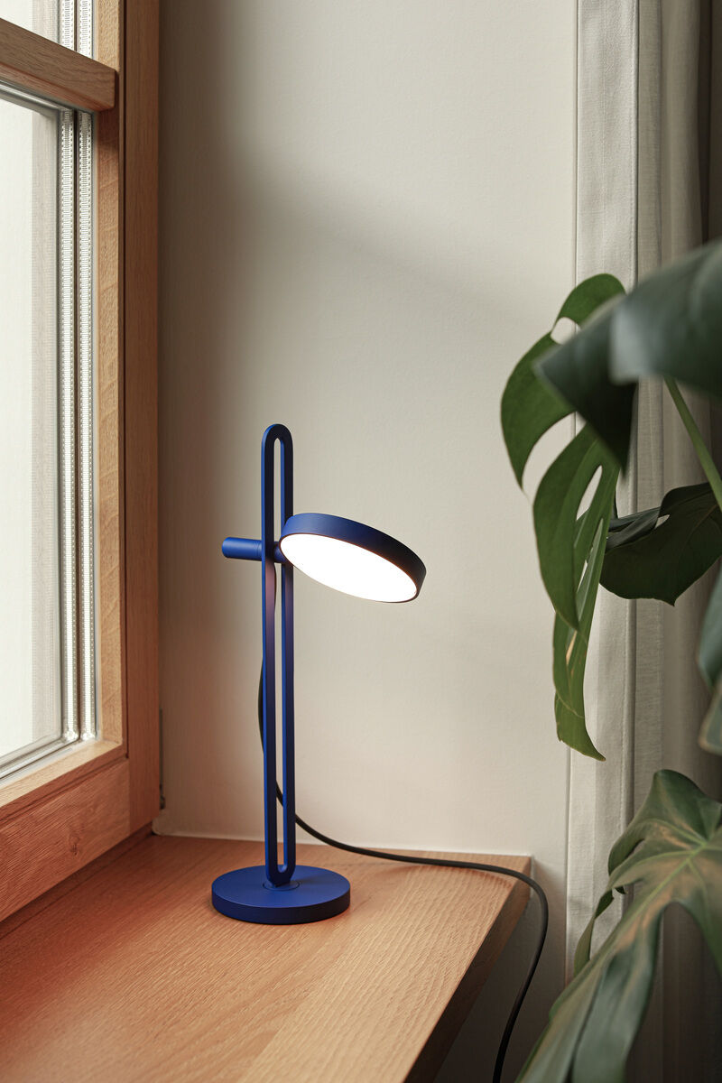 Eine moderne Tischlampe »Echo« mit blauem Körper steht auf einem hellen Holzfensterbrett neben einer grünen Pflanze. Die Lampe hat ein minimalistisches Design und scheint in einem gemütlichen Innenraum gut platziert zu sein.