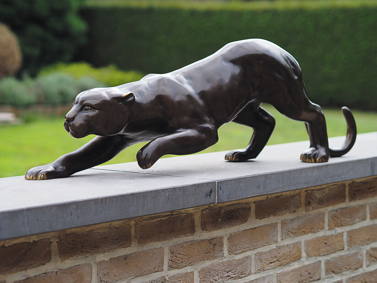 Eine elegante Gartenskulptur eines Panthers. Die Skulptur aus schwarzem Metall zeigt den Panther in einer geschmeidigen, anmutigen Jagdpose. Die Figur steht auf einer gemauerten Gartenmauer. Im Hintergrund ist ein gepflegter Garten mit grüner Hecke und Rasen zu sehen.