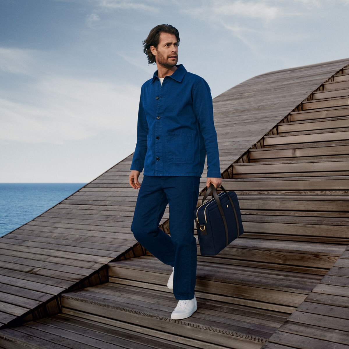 Ein Mann in blauer Kleidung steht auf einer großen Holztreppe mit Blick aufs Meer und hält eine elegante, schwarze Businesstasche der Marke »ENDEAVOUR« von Mismo in der Hand.