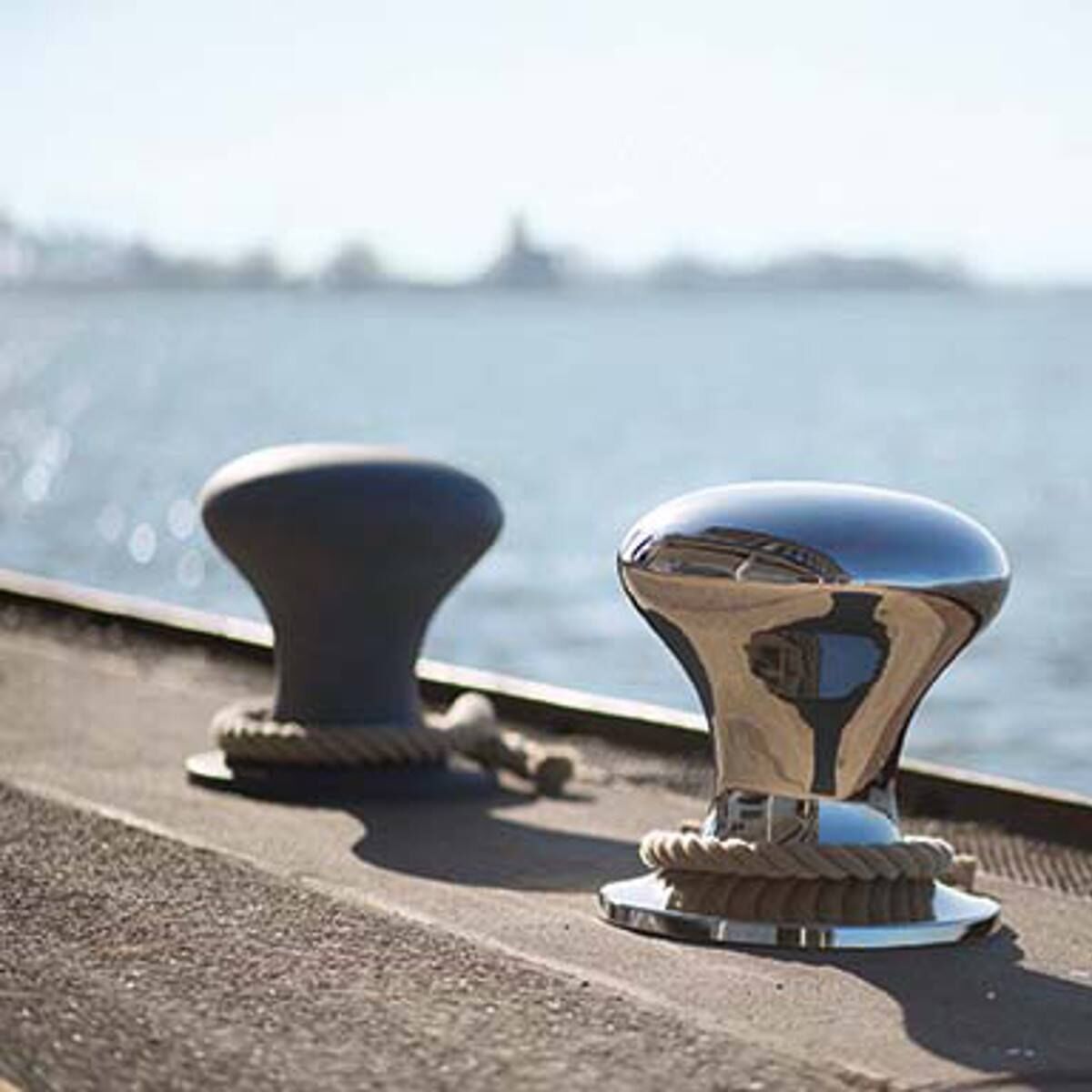 Zwei metallische Hocker mit Segeltau stehen auf einer Steinmauer, mit einem verschwommenen Blick auf das Wasser im Hintergrund. Die Hocker haben eine moderne, glänzende Oberfläche und ein nautisches Design.