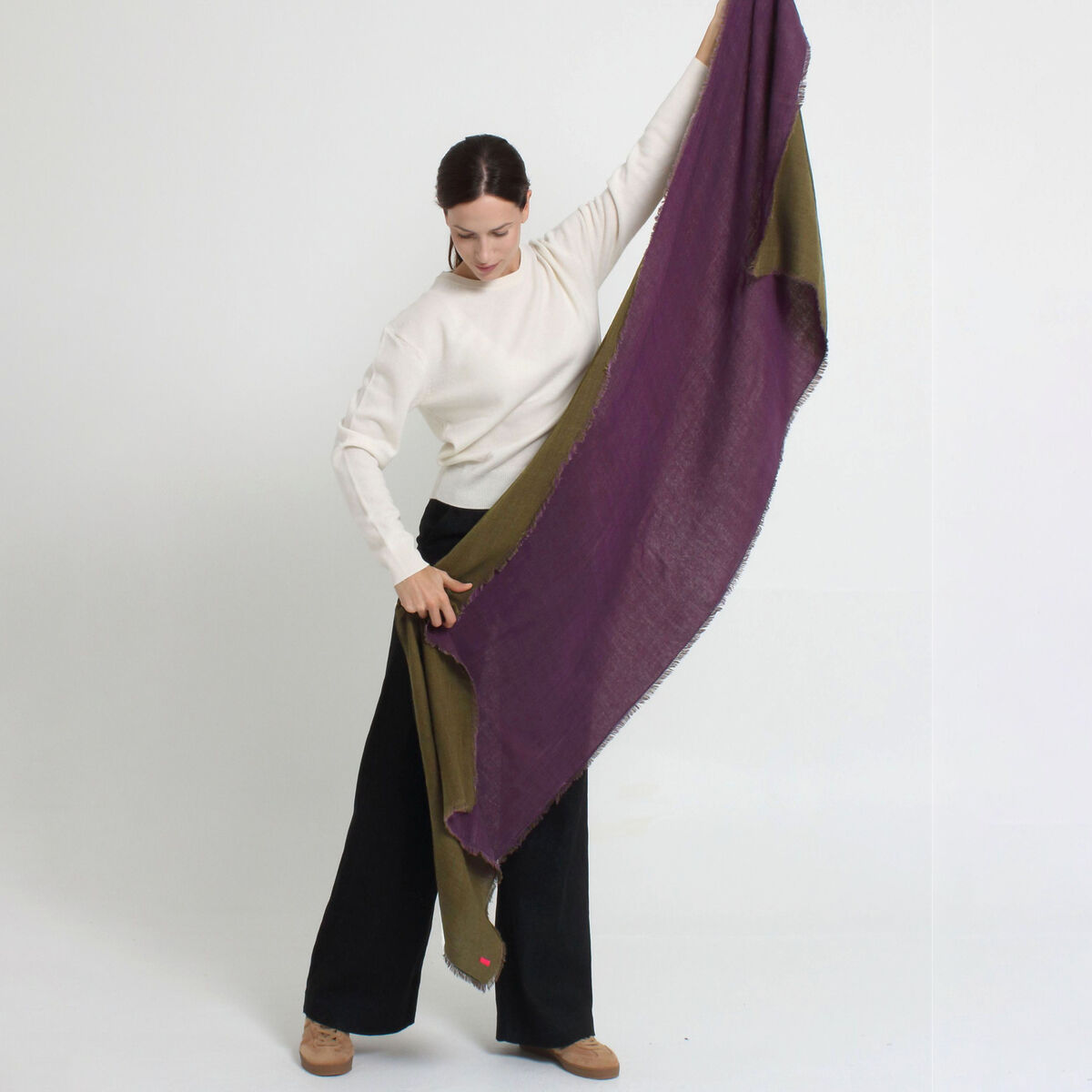 Eine Frau präsentiert den Wollschal »Carla« Violett-Grün von Shalls Barcelona. Der Schal aus 100% Schurwolle hat violette und grüne Flächen und wirkt weich und leicht. Sie trägt dazu einen weißen Pullover und schwarze Hose. Heller Hintergrund, stilvolle Szene.