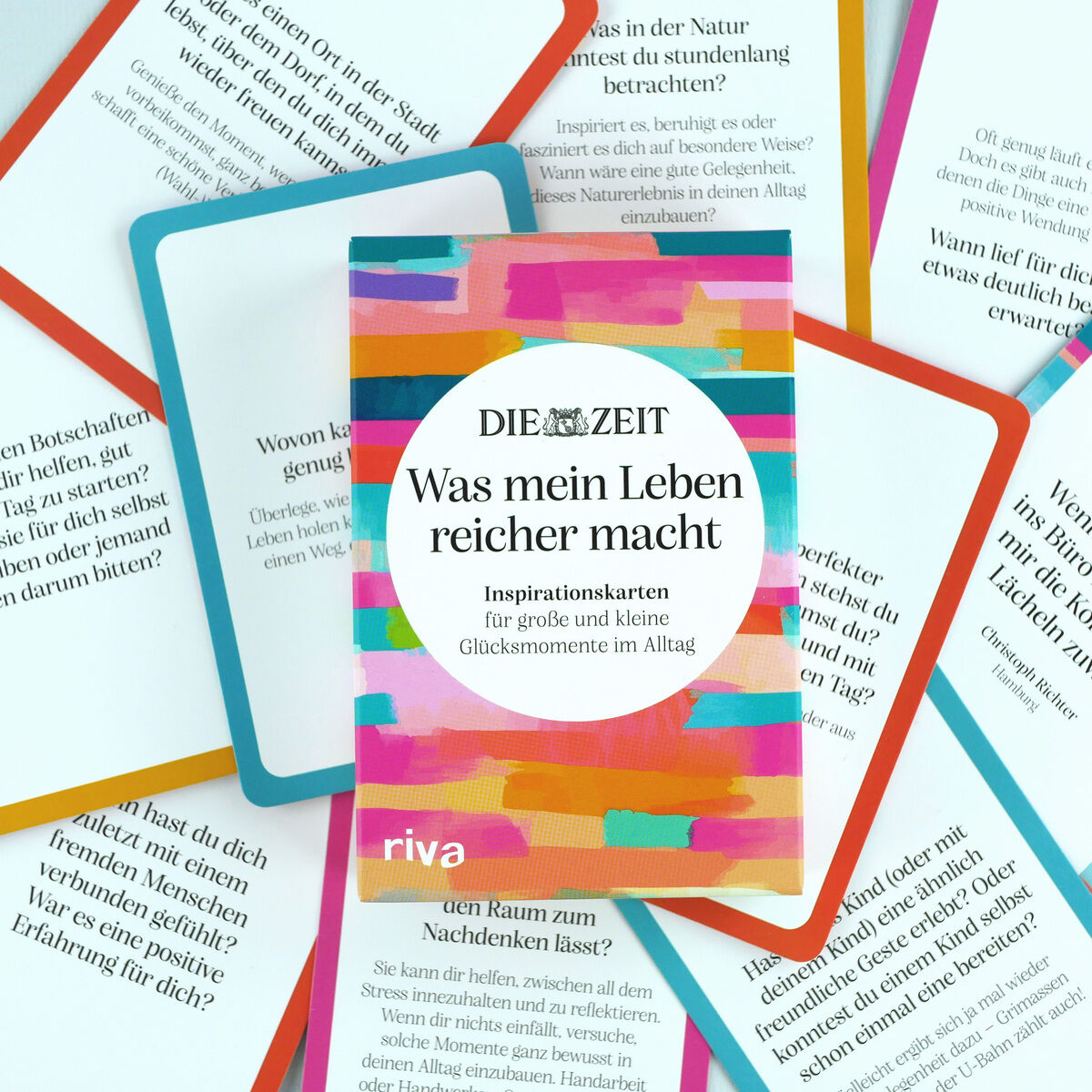 Das Bild zeigt ein Kartenset mit dem Titel „Was mein Leben reicher macht“ von DIE ZEIT/riva Verlag. Die Schachtel ist bunt gemustert, dominieren Pink, Orange und Türkis. Um die Box liegen einzelne Inspirationskarten mit Zitaten und Fragen, gerahmt in kräftigen Farben.
