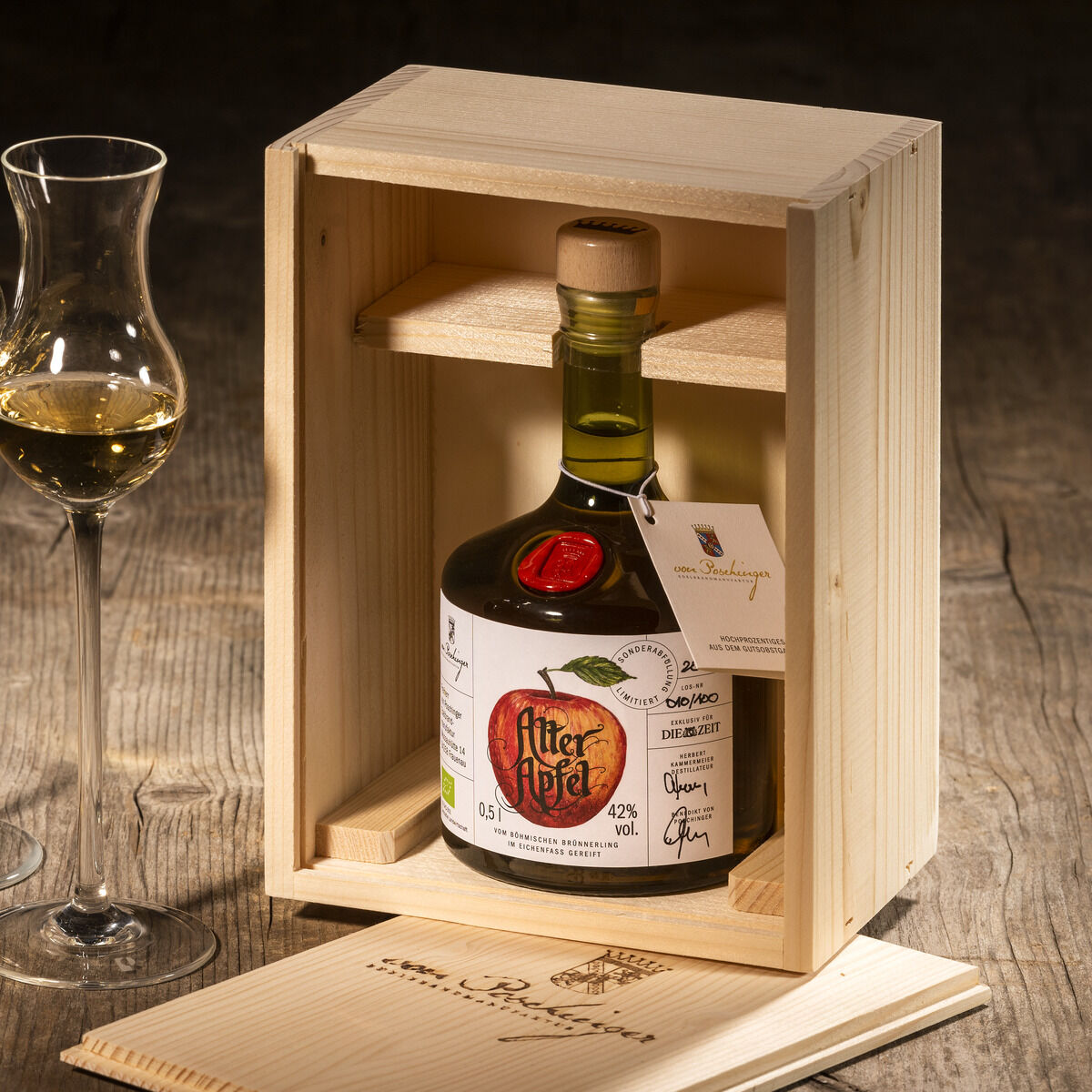 Eine Flasche ZEIT-Sonderedition Edelbrand »Alter Apfel« in einer hellen Holzkiste, begleitet von zwei halb gefüllten Schnapsgläsern und einem Notizbuch auf einem rustikalen Holztisch.