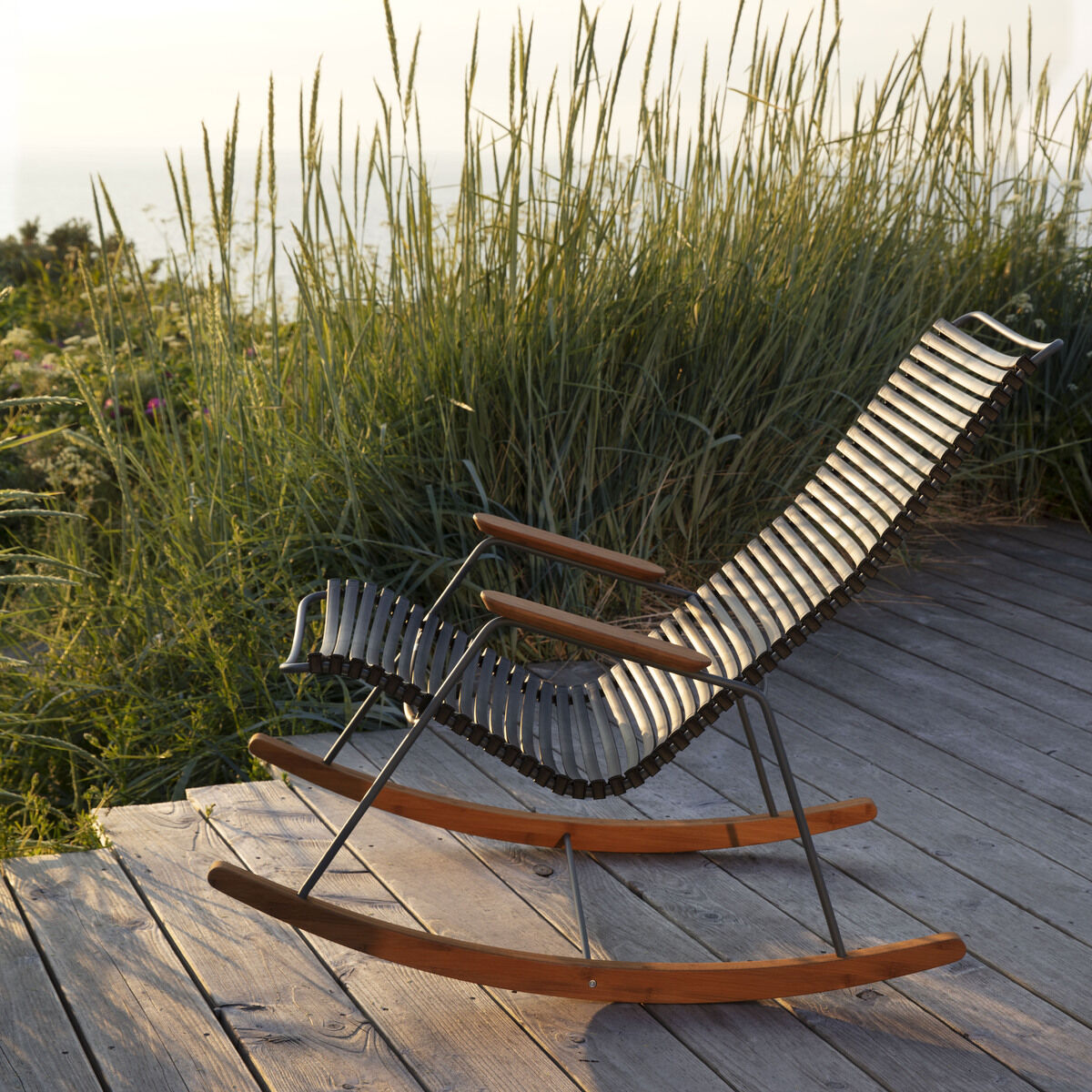 Ein moderner Schaukelstuhl aus Holz und Metalldraht steht auf einem Holzsteg im Freien mit hohem Gras im Hintergrund unter einem sonnigen Himmel.