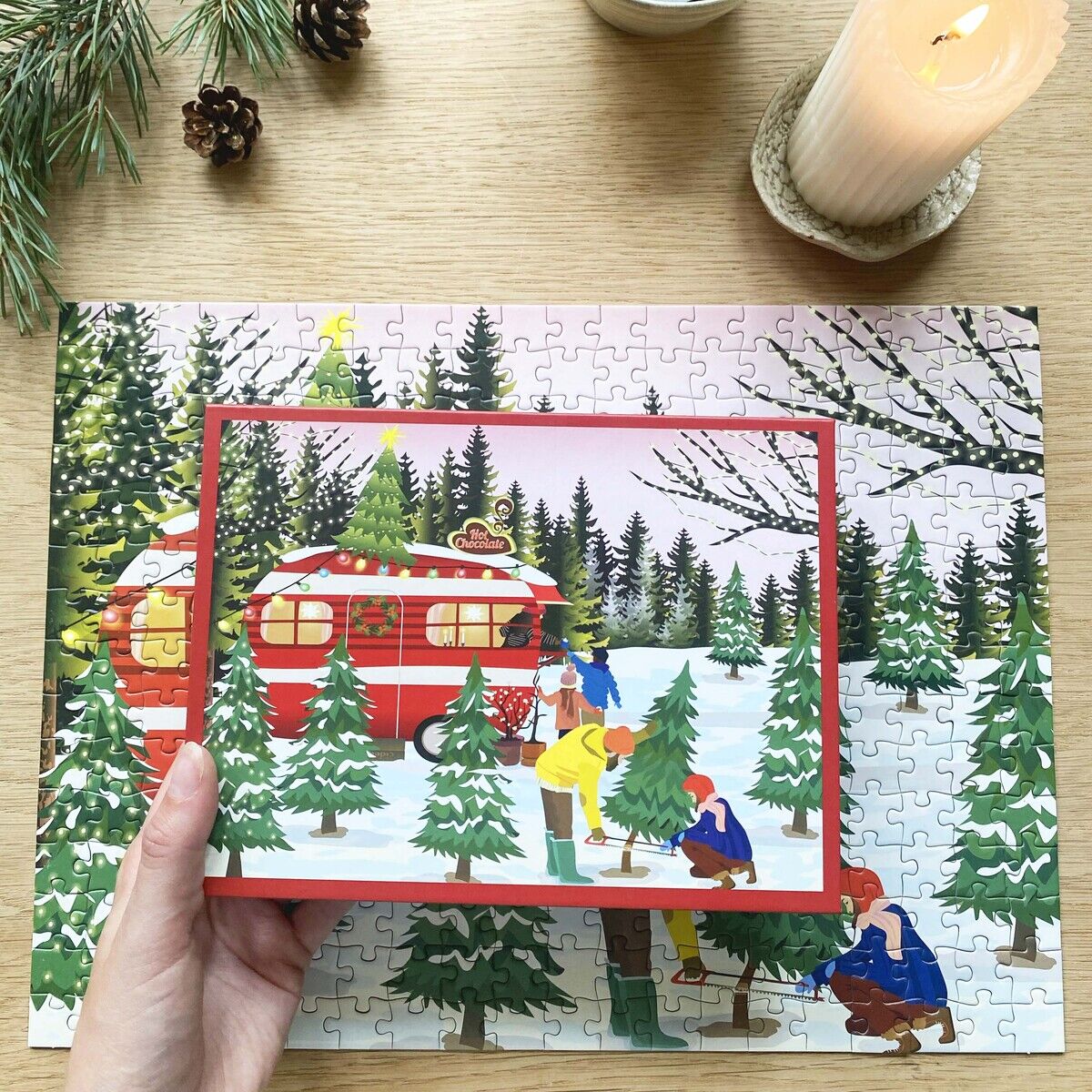 Das Bild zeigt ein 300-Teile-Puzzle „Caravan“ von ViSSEVASSE, das auf einem Holztisch ausgelegt ist. Motive sind ein roter Wohnwagen in winterlicher Landschaft, Tannenbäume und Menschen bei festlicher Aktivität. Dazu stehen eine angezündete Kerze, ein Becher Kaffee und Tannenzweige auf dem Tisch.