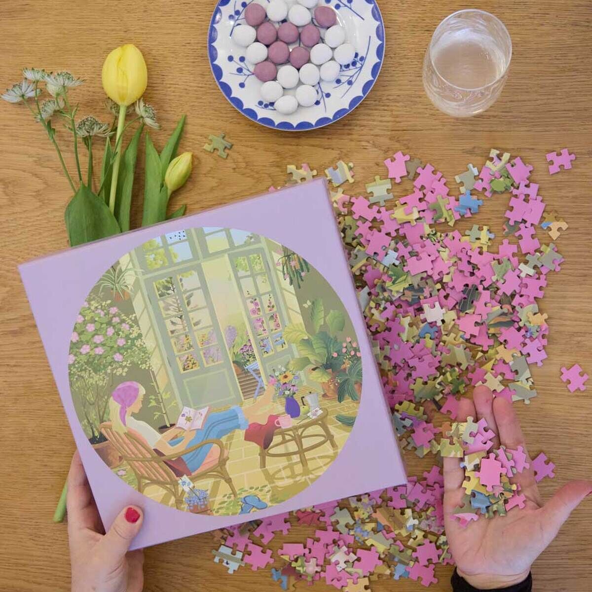 Die Szene zeigt das 1000-Teile Puzzle »Morning Glow« von VISSEVASSE auf Holzoberfläche, mit geöffneter Schachtel, verstreuten Puzzlestücken, Teller mit Süßigkeiten, gelben Tulpen, einem Glas Wasser und Händen, die Teile halten. Fröhliche, ruhige Atmosphäre.