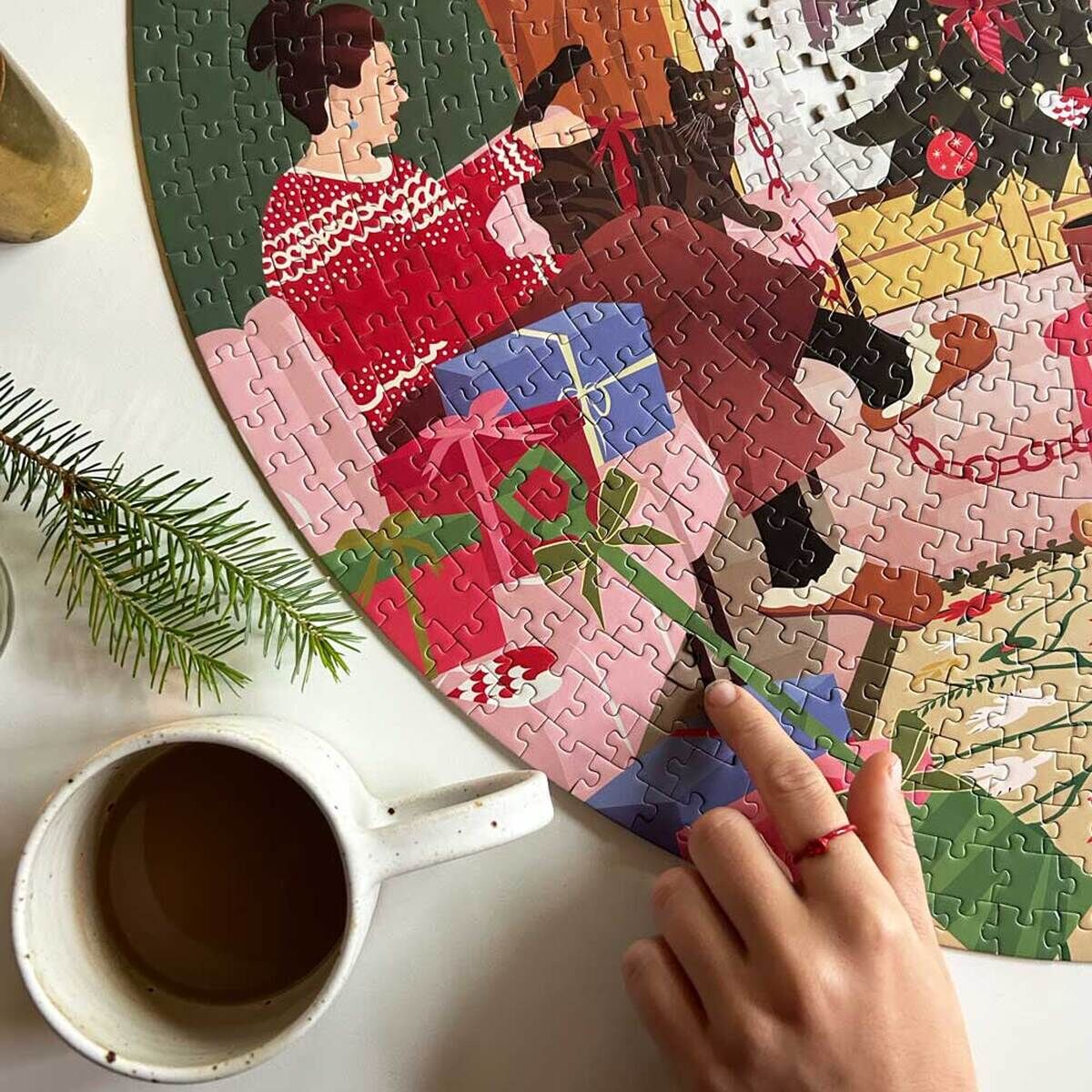 Eine Person sitzt in einem gemütlichen Wohnzimmer neben einem festlich geschmückten Weihnachtsbaum und löst ein Puzzle mit winterlichem Motiv. Neben ihr steht eine Tasse mit heißem Getränk und einige Tannenzweige liegen auf dem Tisch.