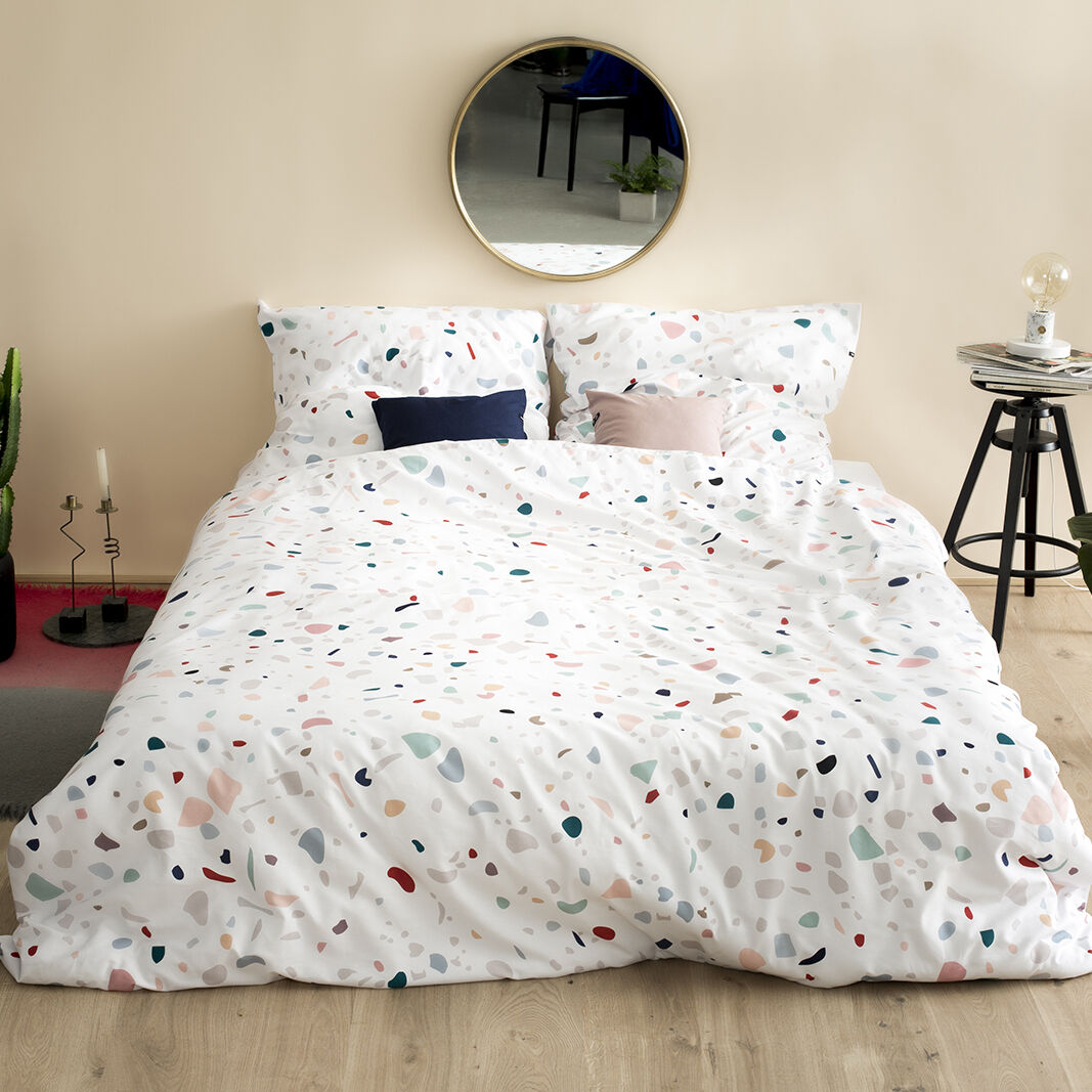 Ein Doppelbett mit Bettwäsche in Terrazzo-Design, umgeben von Pflanzen und moderner Dekoration in einem minimalistisch eingerichteten Schlafzimmer.
