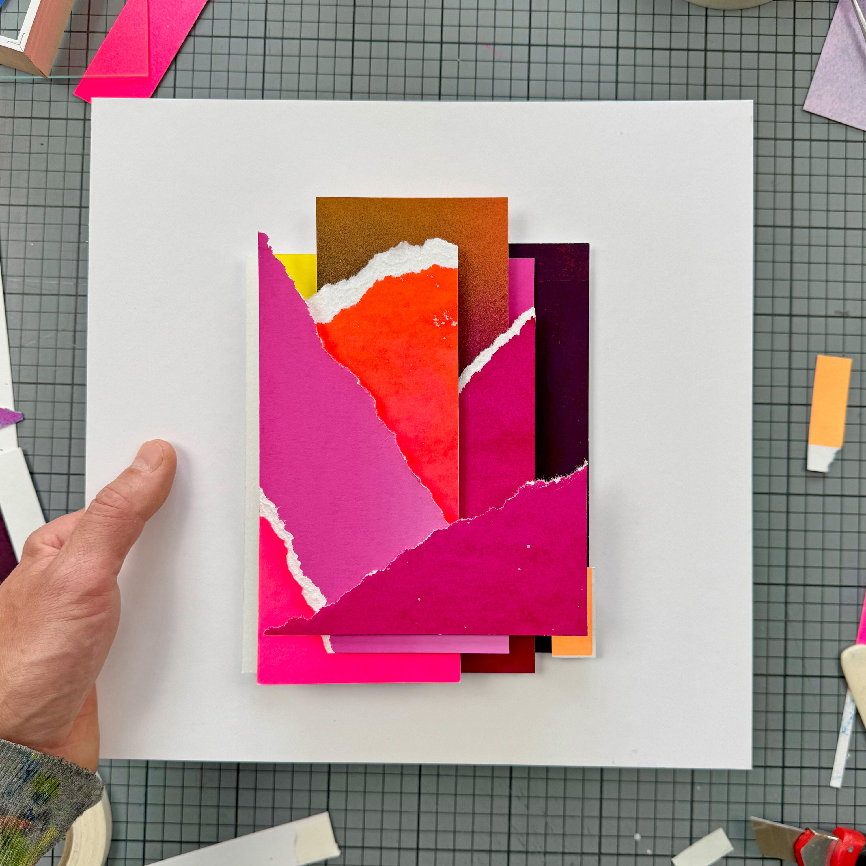 Abstrakte Siebdruck- und Collage-Arbeit mit mehreren übereinandergelegten Papierstücken in leuchtenden Farben wie Pink, Rot, Orange, Gelb, Weiß und Violett auf einem weißen quadratischen Hintergrund. Eine Hand hält das Kunstwerk auf einer karierten Unterlage, im Hintergrund sind weitere farbige Papierschnitte zu sehen.