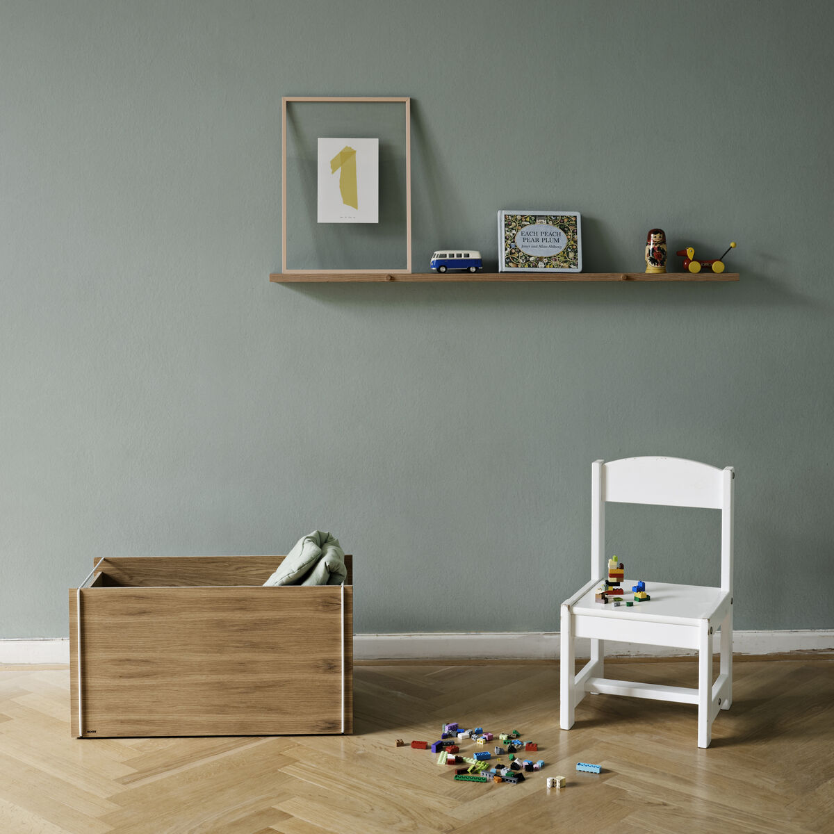 Ein Eichenholz-Wandregal an einer grauen Wand, dekoriert mit einem gerahmten Bild, einem Spielzeugbus und einem kleinen Bilderrahmen. Neben dem Regal steht eine weiße Kindersitzbank und eine Holzspielzeugkiste. Spielzeugteile sind auf dem Hartholzboden verstreut.