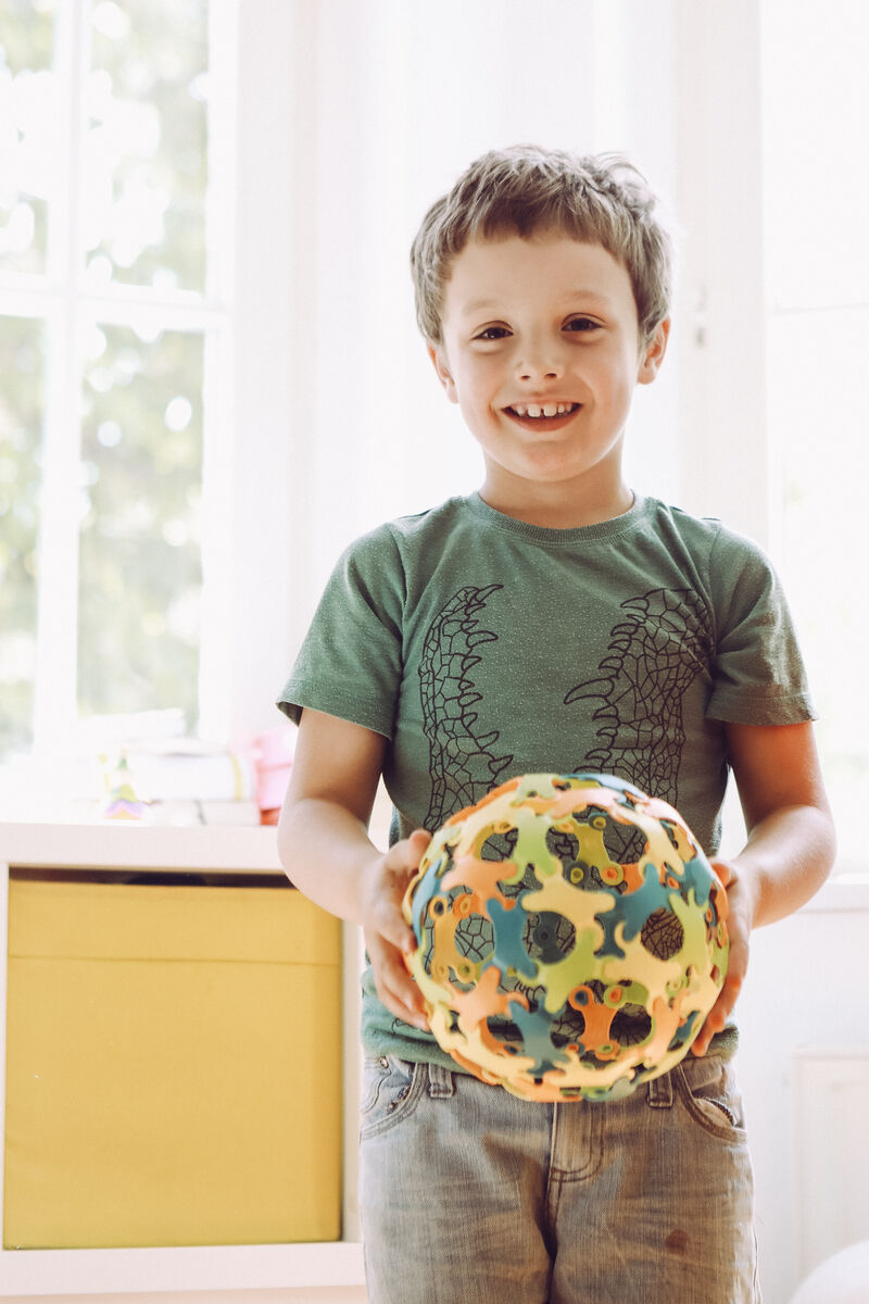 Ein lachendes Kind hält eine bunte Kugel aus miteinander verbundenen Binabo-Teilen. Das Kind trägt ein grünes T-Shirt und steht in einem hellen Raum mit einem gelben Schrank im Hintergrund.