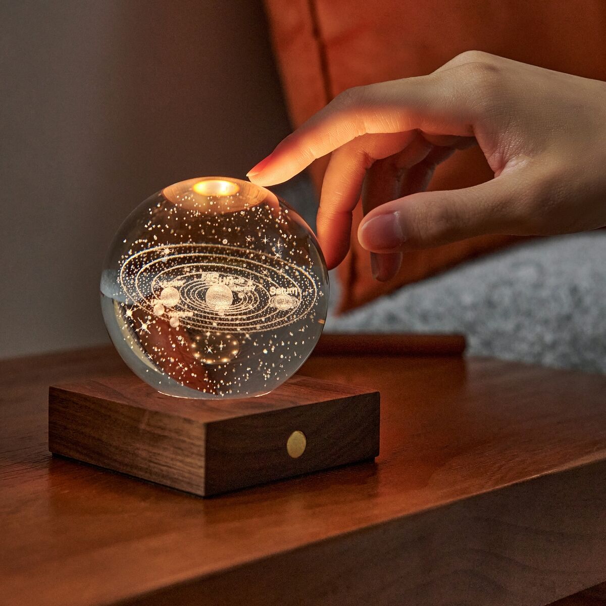 Detailreiche Lampe »Amber Light« von Gingko Design: Eine transparente Kristallkugel mit fein graviertem Sonnensystem sitzt auf einem eleganten, eckigen Sockel aus dunklem Walnussholz. Dimmbares Licht leuchtet atmosphärisch, eine Hand berührt sanft den Kupfer-Touchknopf.