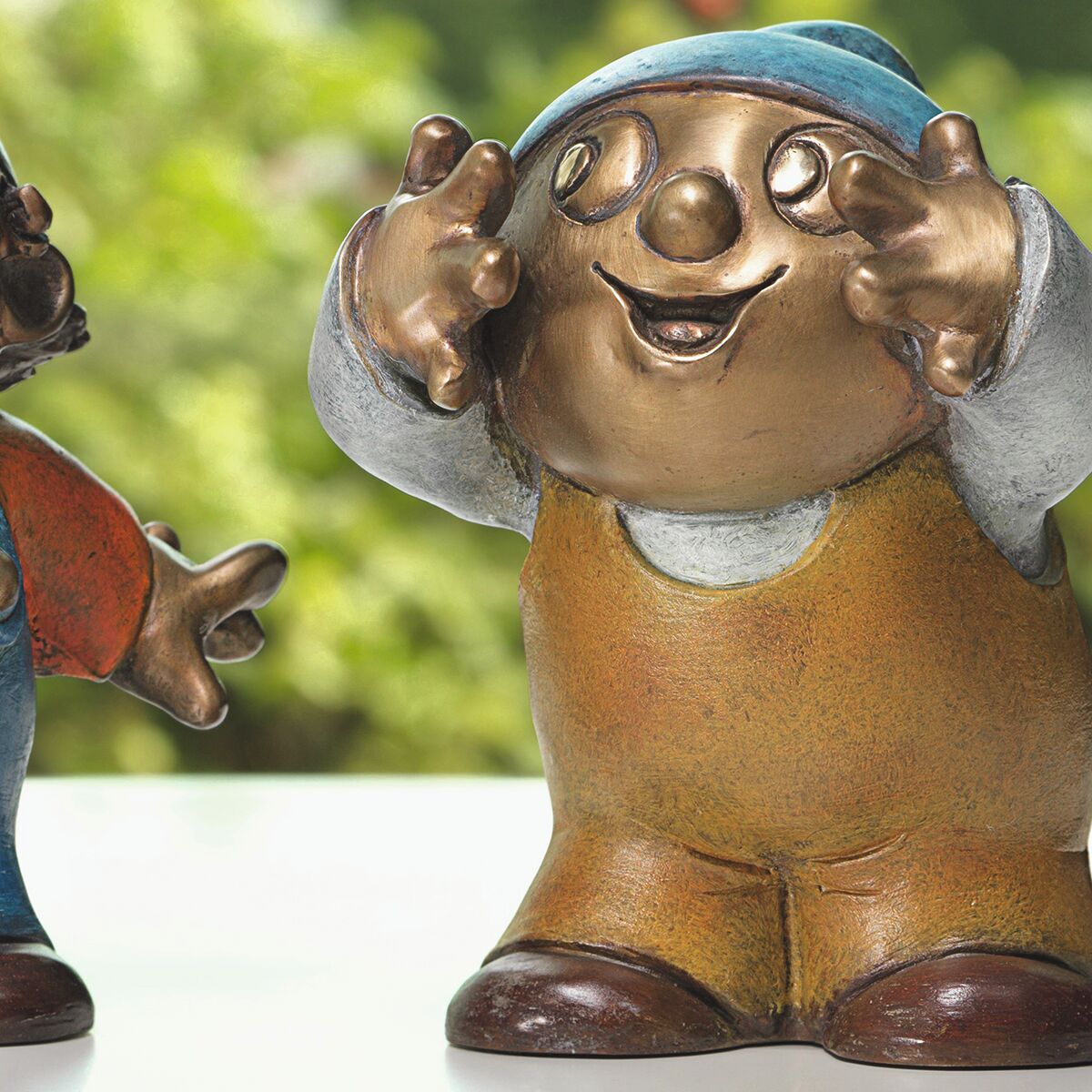 Detailreiche Bronzeskulptur eines Mainzelmännchens mit blauer Mütze, gelbem Overall und fröhlichem Gesichtsausdruck. Die Figur steht aufrecht mit erhobenen Händen, der Hintergrund ist grünlich und unscharf. Kunstvoll handpatiniert, circa 13 cm hoch.