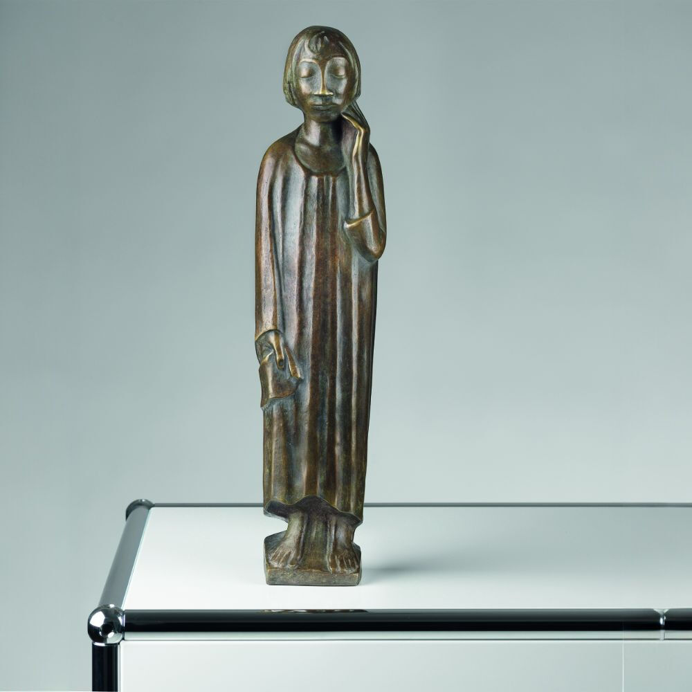 Bronzeskulptur von Ernst Barlach mit dem Titel 'Der Sinnende II' aus dem Jahr 1934. Die Skulptur zeigt eine stehende, nachdenkliche Figur mit der Hand an ihrem Kinn, auf einem modernen Tisch platziert.