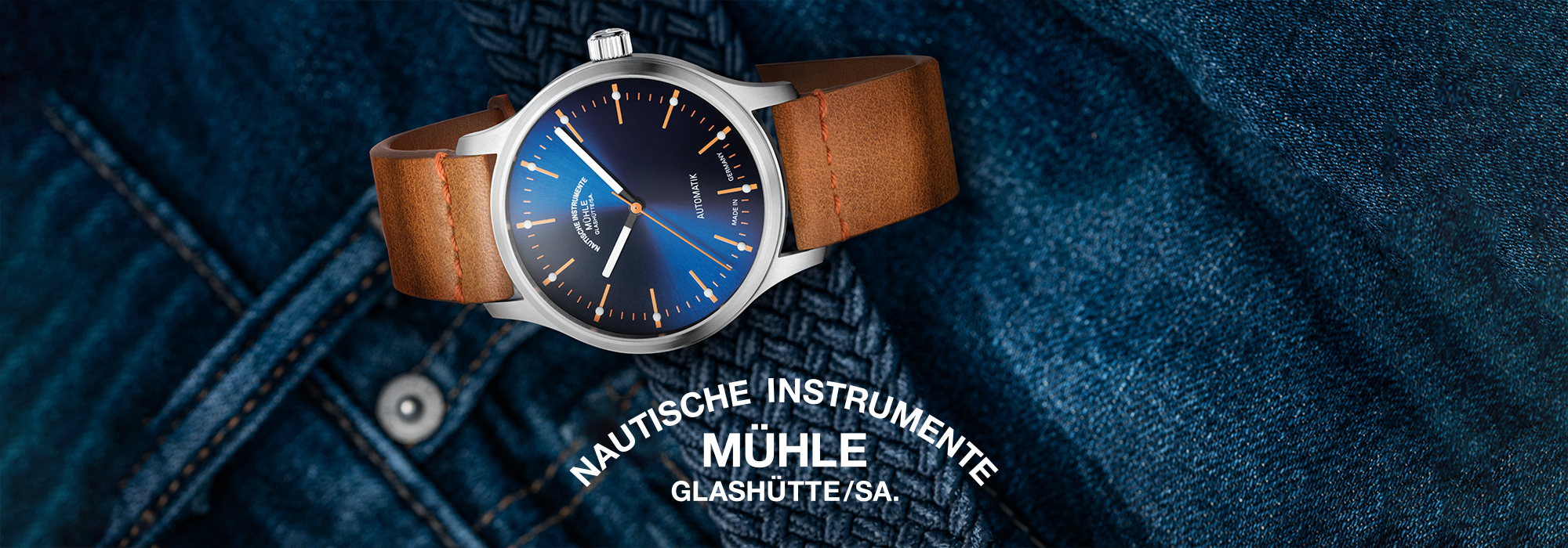 Nahaufnahme einer Mühle Glashütte-Uhr mit blauem Zifferblatt, orangefarbenen Akzenten und einem braunen Lederarmband, das auf Jeansstoff liegt.