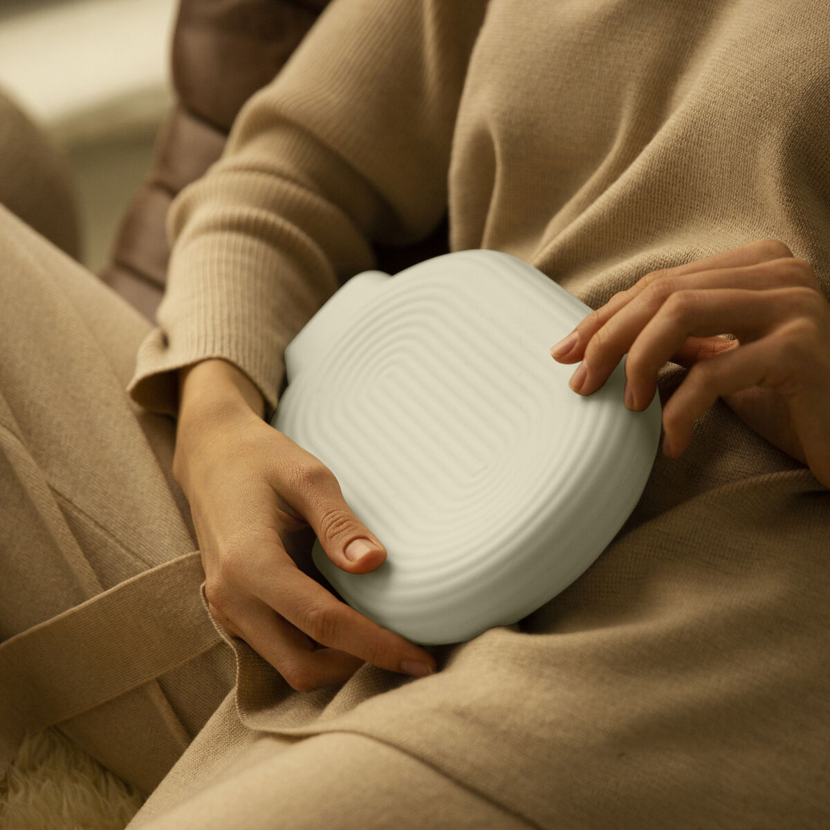 Eine Person sitzt entspannt auf einem beigen Sofa und hält die cremeweiße, elektrische Wärmflasche Silk von Vaerm aus Silikon im Soft-Touch-Design auf dem Bauch. Umgeben von weichen Kissen verströmt die Szene beruhigende Wärme und Komfort.