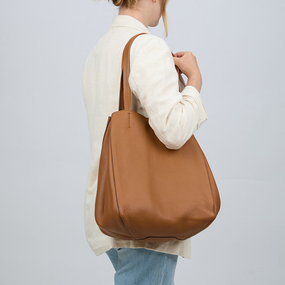 Eine Frau trägt die ZEIT-Tasche »Cep« der Marke Guerer & Guerer in Cognac auf der Schulter. Die große, cognacfarbene Ledertasche besticht durch ihr schlichtes, hochwertiges Design und feine Narbung. Sie trägt einen beigen Blazer und eine hellblaue Jeans.