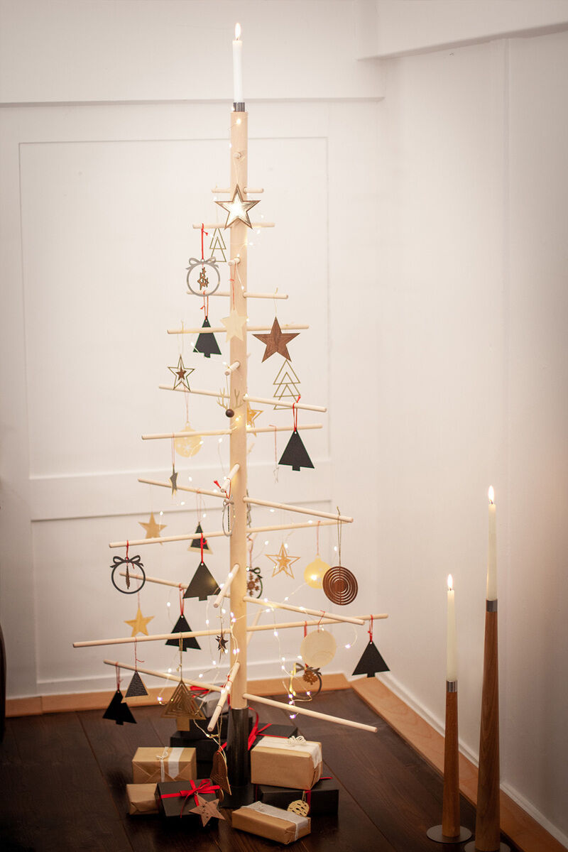 Ein abstrakter Weihnachtsbaum mit minimalistischen Design, aus Holz gefertigt und mit Stahlständer. Er ist mit gelblichen Lichtern und verschiedenen Anhängern in Schwarz, Gold und Rot geschmückt. Rundherum befinden sich verpackte Geschenke. Im Hintergrund ist ein weißes Zimmer mit Holzboden zu sehen, an zwei Kerzenständern brennen Kerzen.