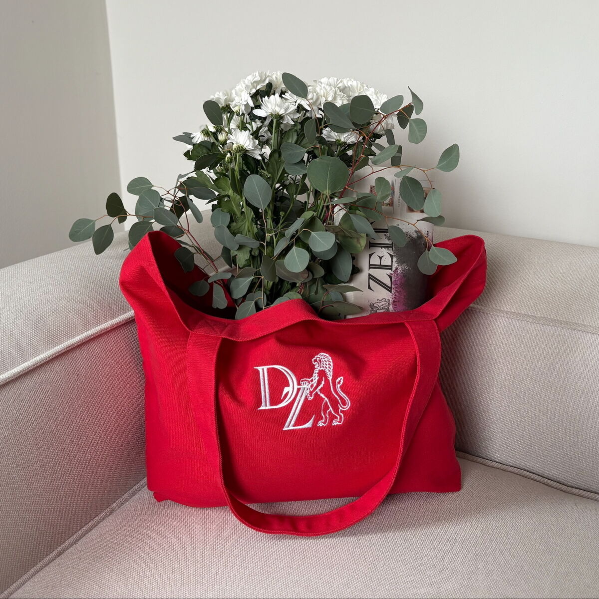 Eine korallenrote Canvas-Tasche von ZEIT befindet sich auf einem beigen Sofa. Die Tasche enthält weiße Blumen und eine Zeitung. Das Design ist minimalistisch und die Tasche hat einen umweltfreundlichen Stil.