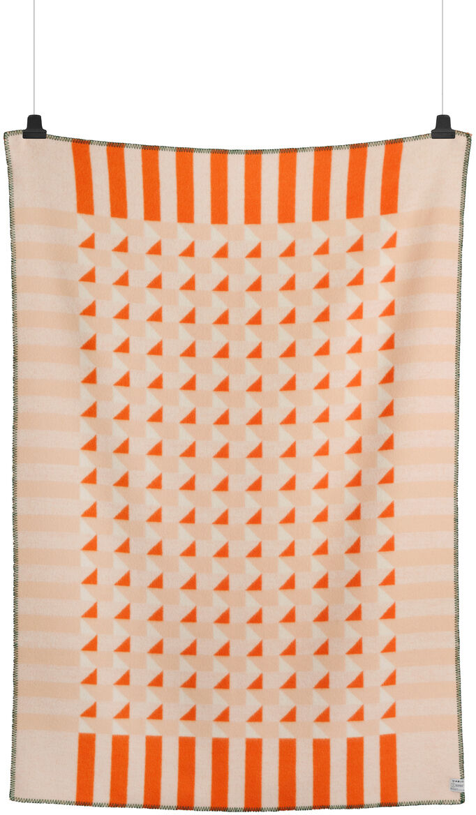 Eine Lammwolldecke mit dem Namen »KVAM« hängt vertikal aufgehängt. Die Decke zeigt ein modernes Design mit einem abstrakten Muster aus weißen und orangefarbenen Dreiecken und Streifen. Der Rand ist schwarz eingefasst.