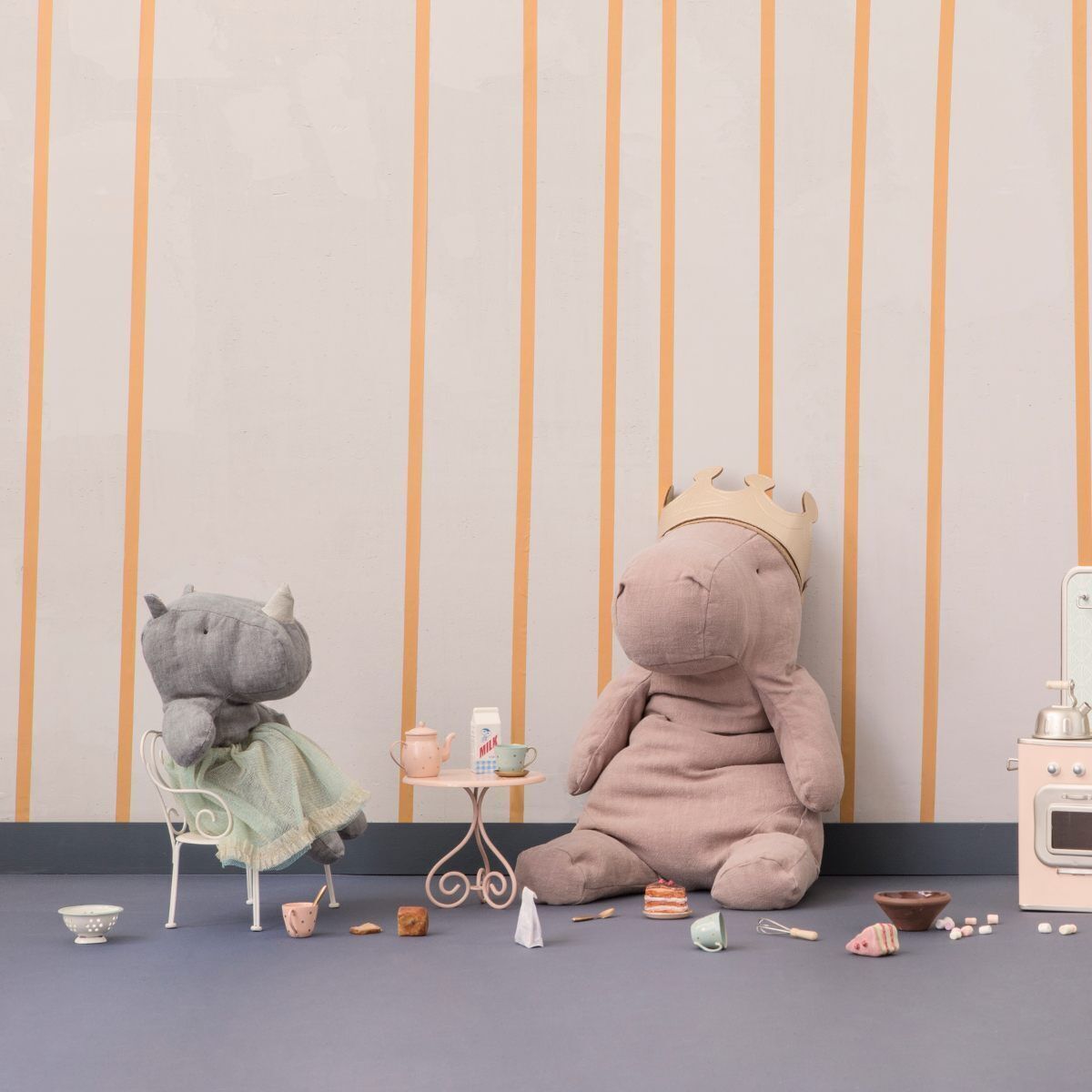 Zwei plüschige Nashorn-Kuscheltiere, eines auf einem weißen Stuhl und eines auf dem Boden; das auf dem Boden sitzt und eine Krone trägt. Im Hintergrund ist eine cremefarbene Wand mit gelben senkrechten Streifen zu sehen. Einige kleine Spielzeugtassen und Dekorationen sind ebenfalls im Bild.