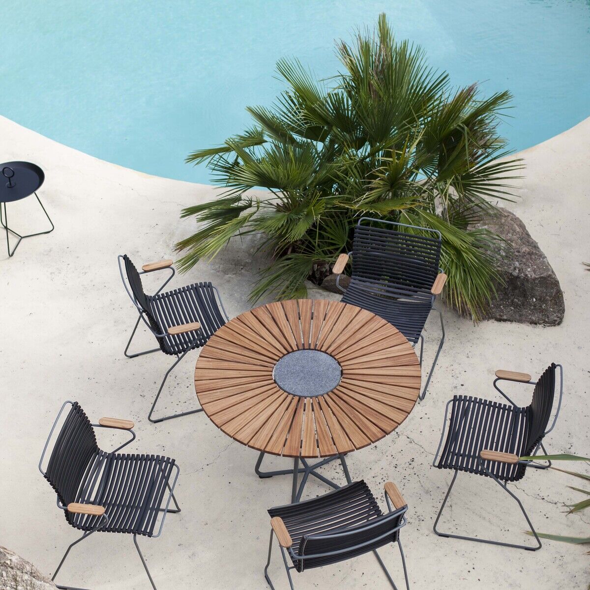 Gartenmöbel-Set um einen runden Tisch aus Holz und Metall, bestehend aus sechs schwarzen »CLICK Dining Chairs« mit Armlehnen. Arrangement befindet sich auf einer weißen Oberfläche inmitten von Palmen und neben einem Swimmingpool.