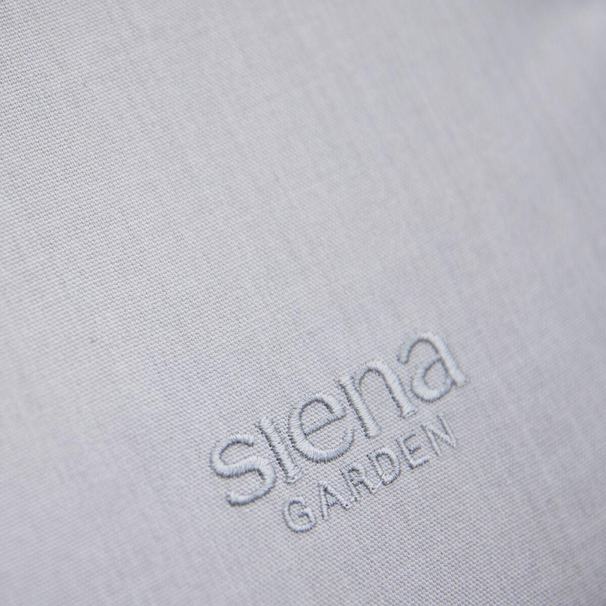 Nahaufnahme eines hellgrauen Stoffes mit gesticktem 'siena GARDEN' Logo, das zu einer hochwertigen Swingliege der Marke Aliano gehört.