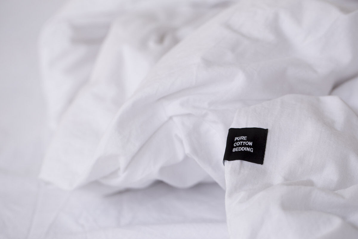 Nahaufnahme von weißer Bettwäsche »Pure« aus 100% Baumwolle mit einem schwarzen Etikett mit der Aufschrift 'PURE COTTON BEDDING'.