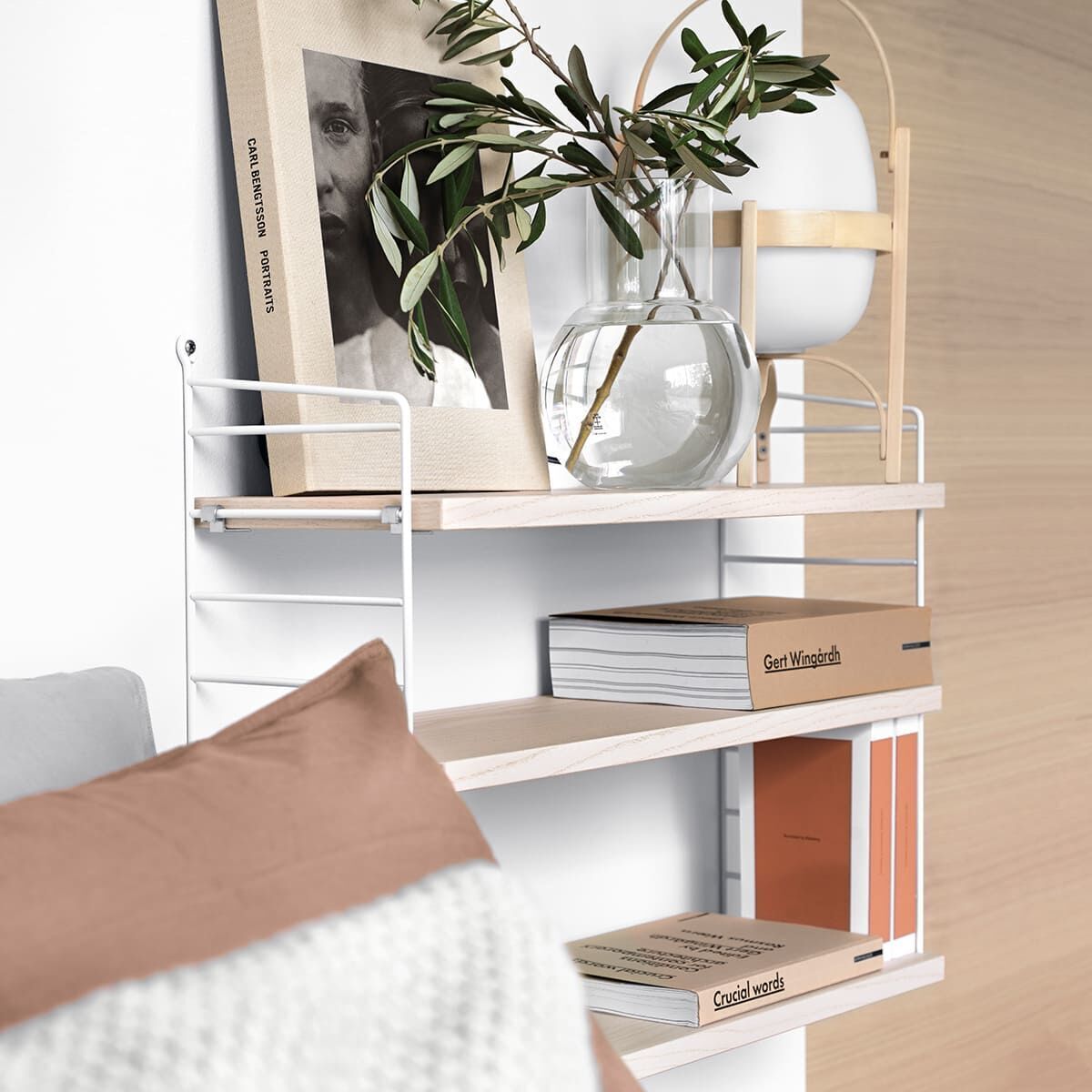 Ein minimalistisches Wandregal »String Pocket Regal« mit drei Holzböden ist an einer weißen Wand montiert. Auf dem oberen Regalbrett stehen gerahmte Bilder und eine Vase mit grünen Zweigen. Auf den unteren beiden Regalbrettern befinden sich Bücher. Im Vordergrund ist ein Teil eines Sofas mit einem beigen Kissen zu sehen.