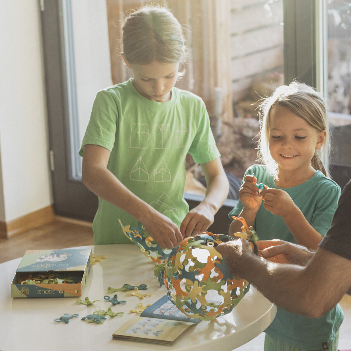 Ein Vater und zwei Kinder spielen gemeinsam mit einem Spiel namens 'Binabo', das aus 60 Teilen besteht. Sie sitzen an einem Tisch und bauen eine konstruktion mit verschiedenfarbigen Teilen.