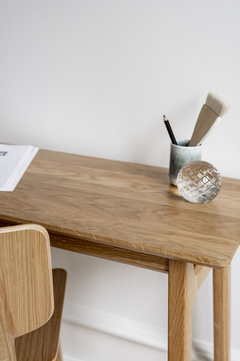 Ein Schreibtisch »Freya« aus Eiche, fotografiert in einer minimalistischen Umgebung. Der Tisch zeigt eine glatte, natürliche Holzstruktur und wird von einem passenden Stuhl begleitet. Auf dem Tisch stehen ein Behälter mit Stiften und Pinseln sowie eine dekorative Kugel. Im Hintergrund ist eine weiße Wand zu sehen.