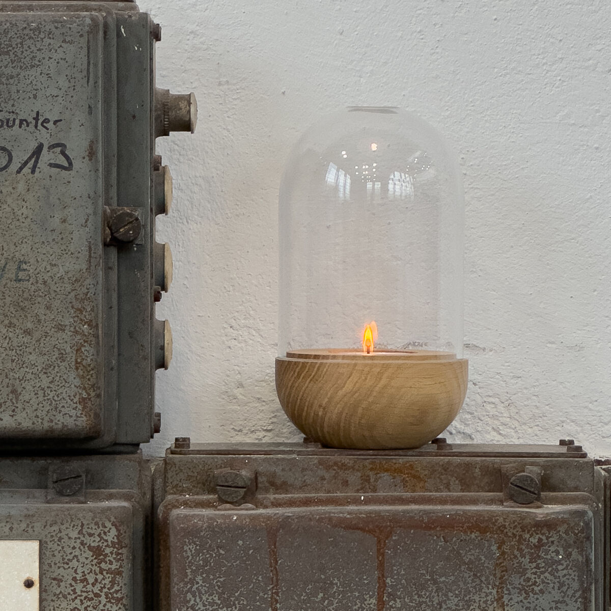 Auf einem alten, grauen Metallschaltkasten steht ein edles Windlicht »GLOW« von finbeaux. Es besteht aus einer massiven Eichenschale und einem hohen, mundgeblasenen Klarglashaube. Im Innern brennt eine kleine Flamme. Die Wand wirkt leicht strukturiert und weiß.