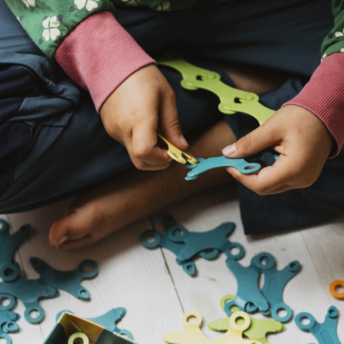 Ein Kind sitzt auf dem Boden und spielt mit farbigen Teilen des Spiels 'Binabo'. Die Teile sind hauptsächlich grün, blau und gelb.