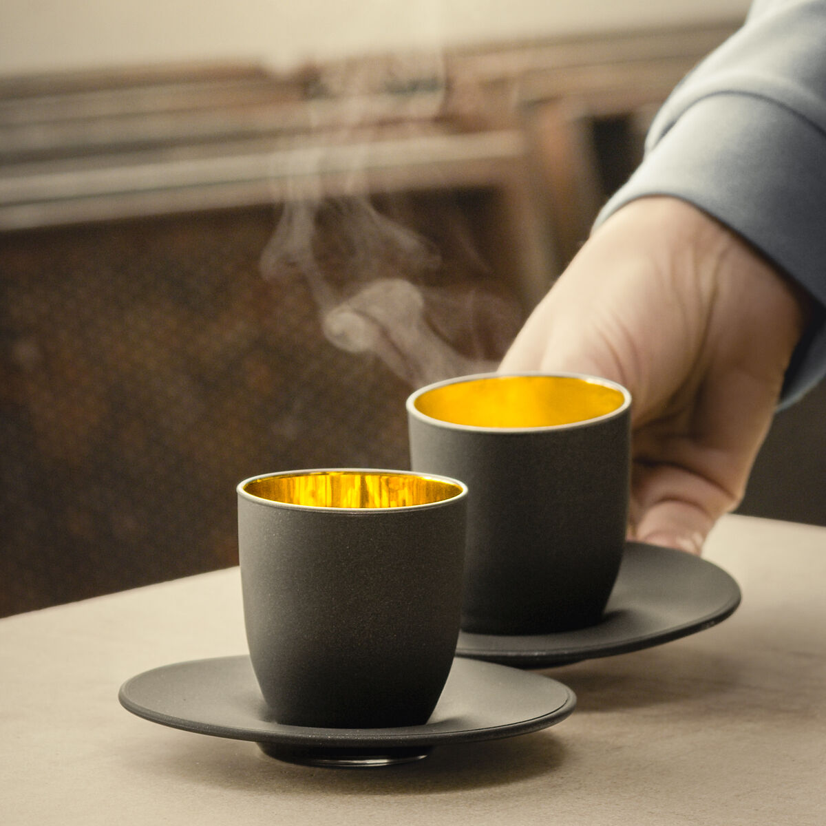 Zwei schwarze Espressobecher mit goldenem Innenrand stehen auf Untertellern. Aus den Tassen steigt Dampf auf. Eine Hand serviert die Tassen auf einer beigen Oberfläche in gemütlicher Atmosphäre.
