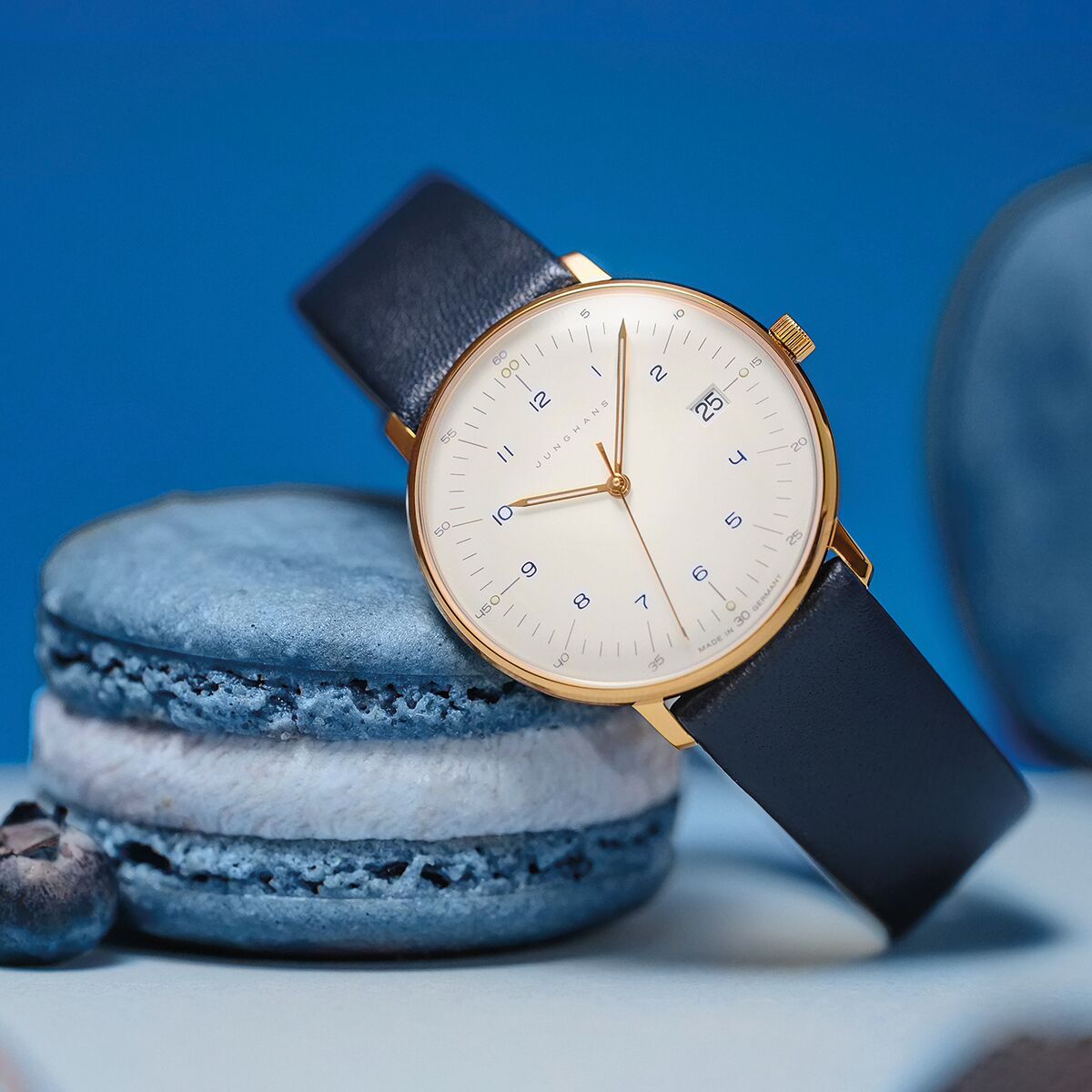 Auf dem Bild ist die ZEIT Sonderedition »max bill Damen« Uhr von Junghans zu sehen. Die Uhr besticht durch ein weißes Zifferblatt mit goldfarbenem Gehäuse und blauen Lederarmband. Die Uhr liegt auf zwei blauen Macarons, im Hintergrund unscharfe blaue Elemente.
