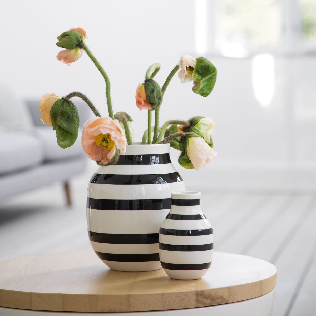 Zwei gestreifte Vasen, eine größere und eine kleinere, stehen auf einem hellen Holztisch. Die größere Vase enthält stielige Blumen mit grünen Blättern und rosa Blüten. Der Hintergrund ist weich und verschwommen, einschließlich eines Fensters mit Tageslicht und Teilen eines Sofas.