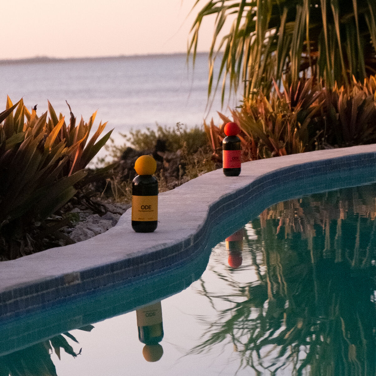 Zwei Aperitifflaschen, eine gelbe und eine rote, stehen auf dem Rand eines Swimmingpools mit tropischen Pflanzen im Hintergrund und einem See oder Meer im Vordergrund bei Sonnenuntergang.